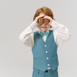 A young boy in a teal suit and bow tie holds two round objects to his eyes, mimicking binoculars, against a plain light background.