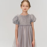 A young girl stands against a white background, wearing a light purple tulle dress with puffed short sleeves and KIKI and JOJO Girls Silver Bow Hair Clips (Set of 2) that add subtle decorative details to her look.