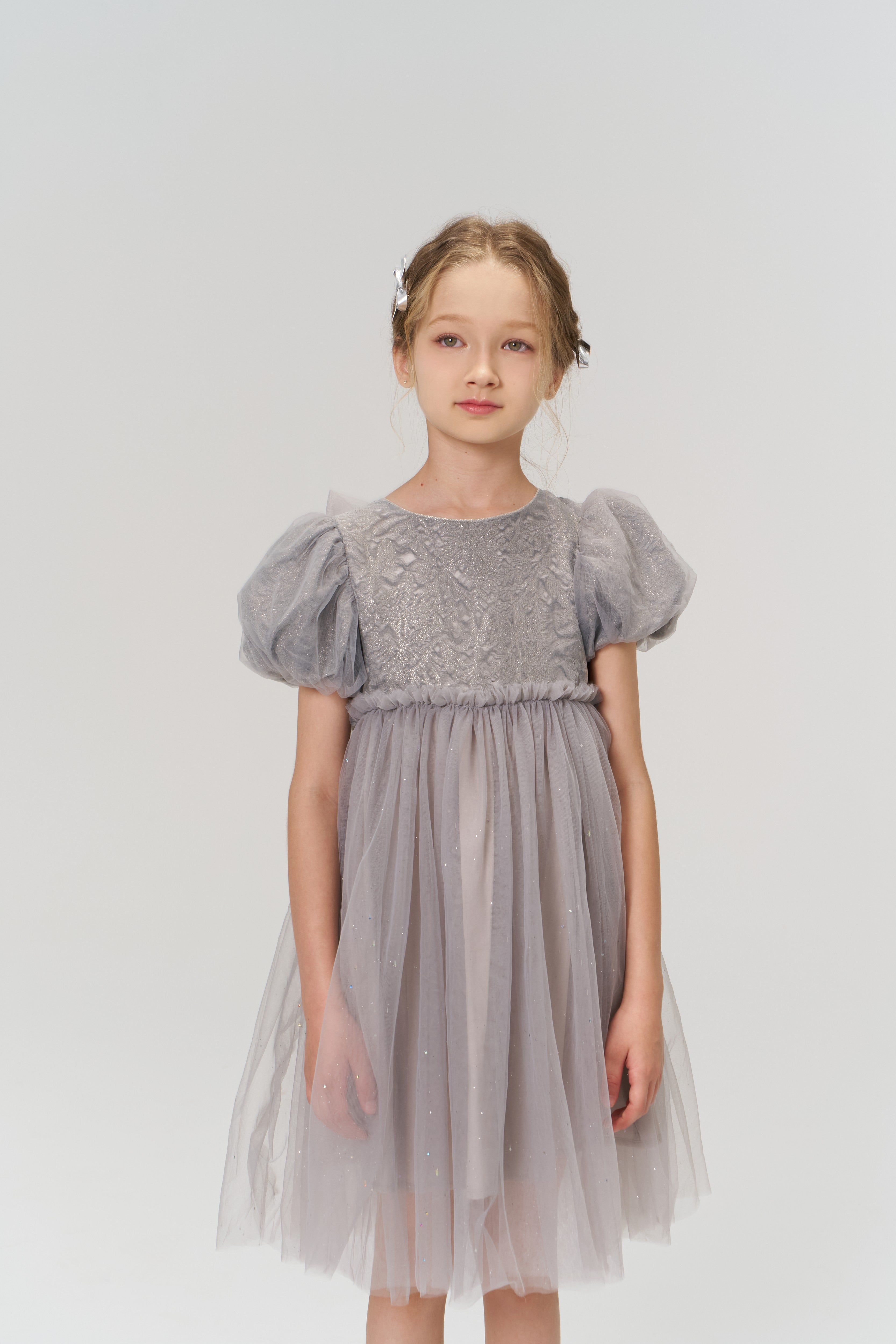 A young girl stands against a white background, wearing a light purple tulle dress with puffed short sleeves and KIKI and JOJO Girls Silver Bow Hair Clips (Set of 2) that add subtle decorative details to her look.