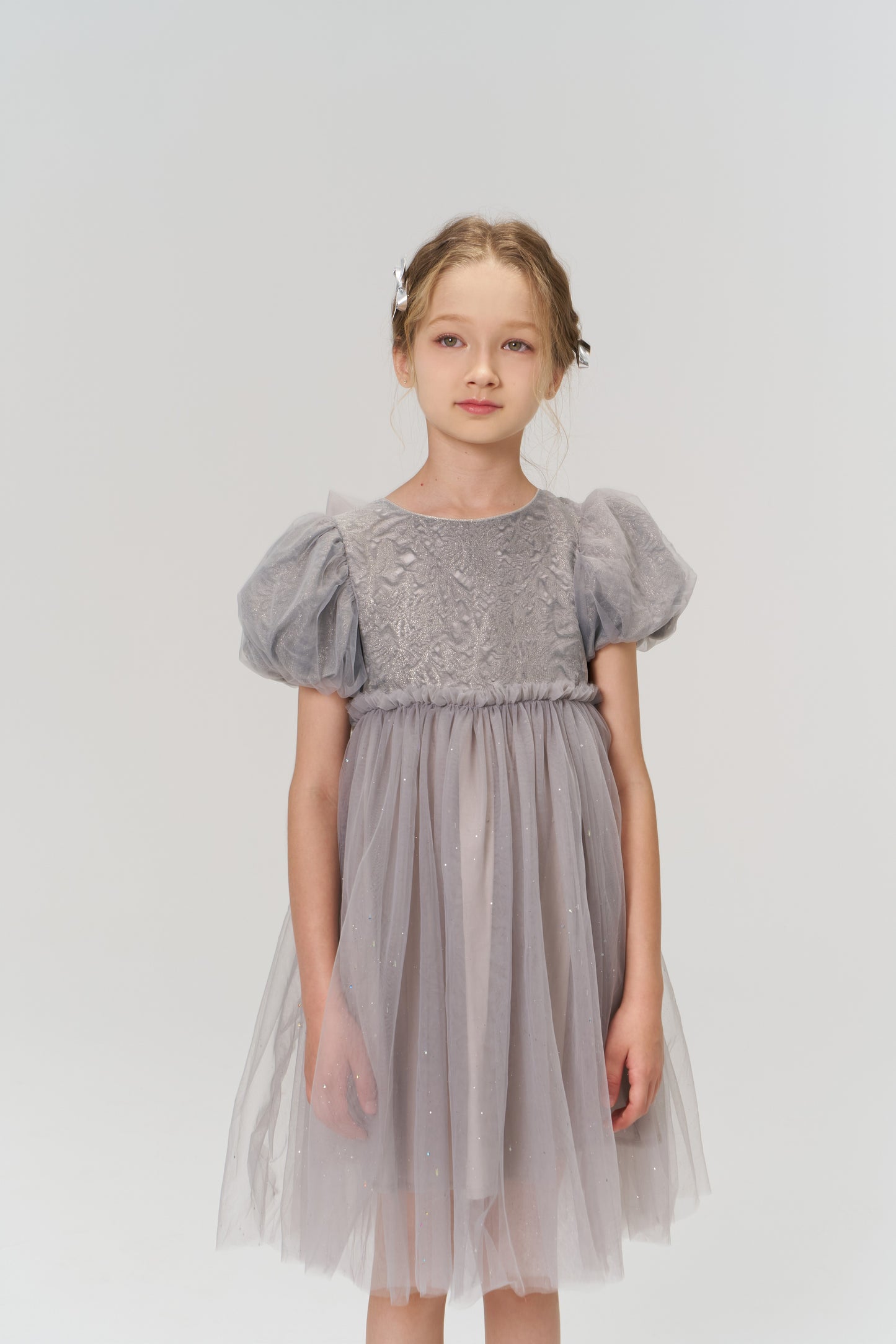 A young girl stands against a white background, wearing a light purple tulle dress with puffed short sleeves and KIKI and JOJO Girls Silver Bow Hair Clips (Set of 2) that add subtle decorative details to her look.
