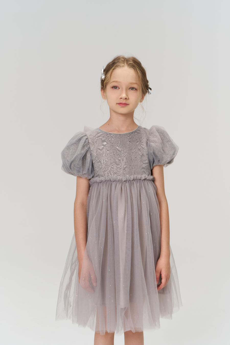 A young girl stands against a white background, wearing a light purple tulle dress with puffed short sleeves and KIKI and JOJO Girls Silver Bow Hair Clips (Set of 2) that add subtle decorative details to her look.