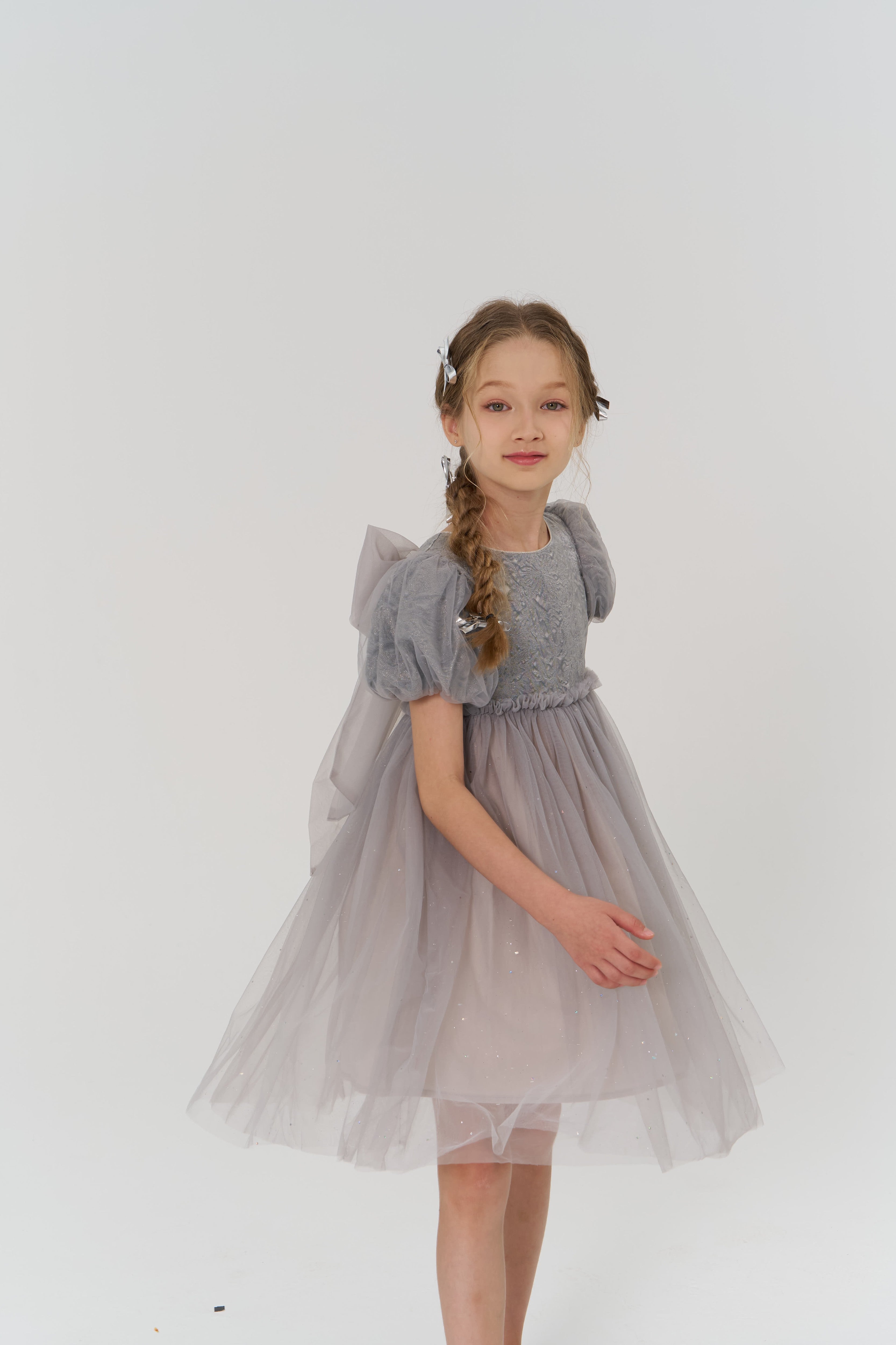 A young girl with long braided hair adorned with KIKI and JOJO’s Girls Silver Bow Hair Clips (set of 2) wears a light purple tulle dress with puffed sleeves and a large back bow, standing against a plain white background.