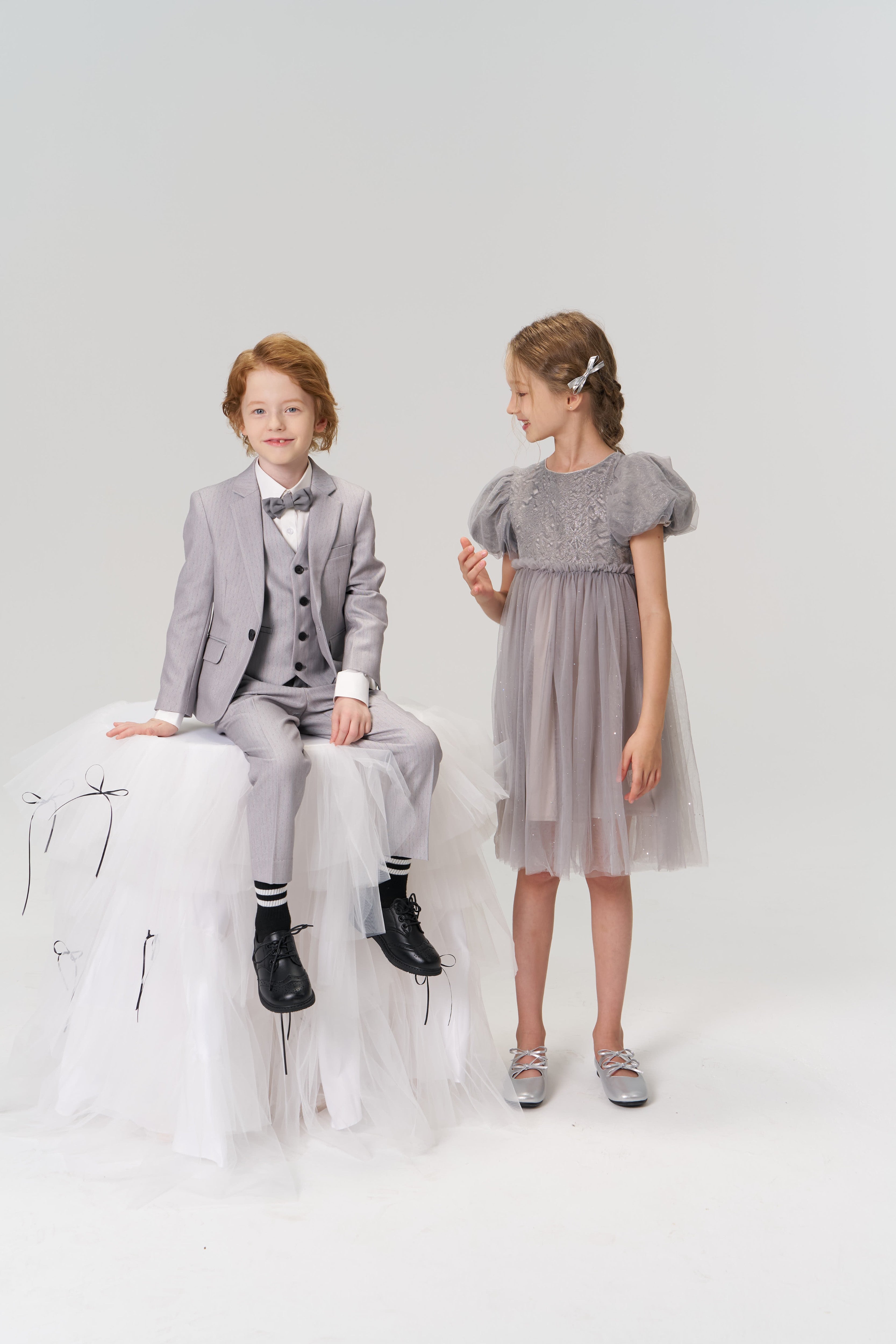 A young girl wearing KIKI and JOJO Girls Silver Bow Hair Clips stands in a gray dress next to a boy in a light gray suit, who sits on a white tulle-covered stool, both posed against a plain background.