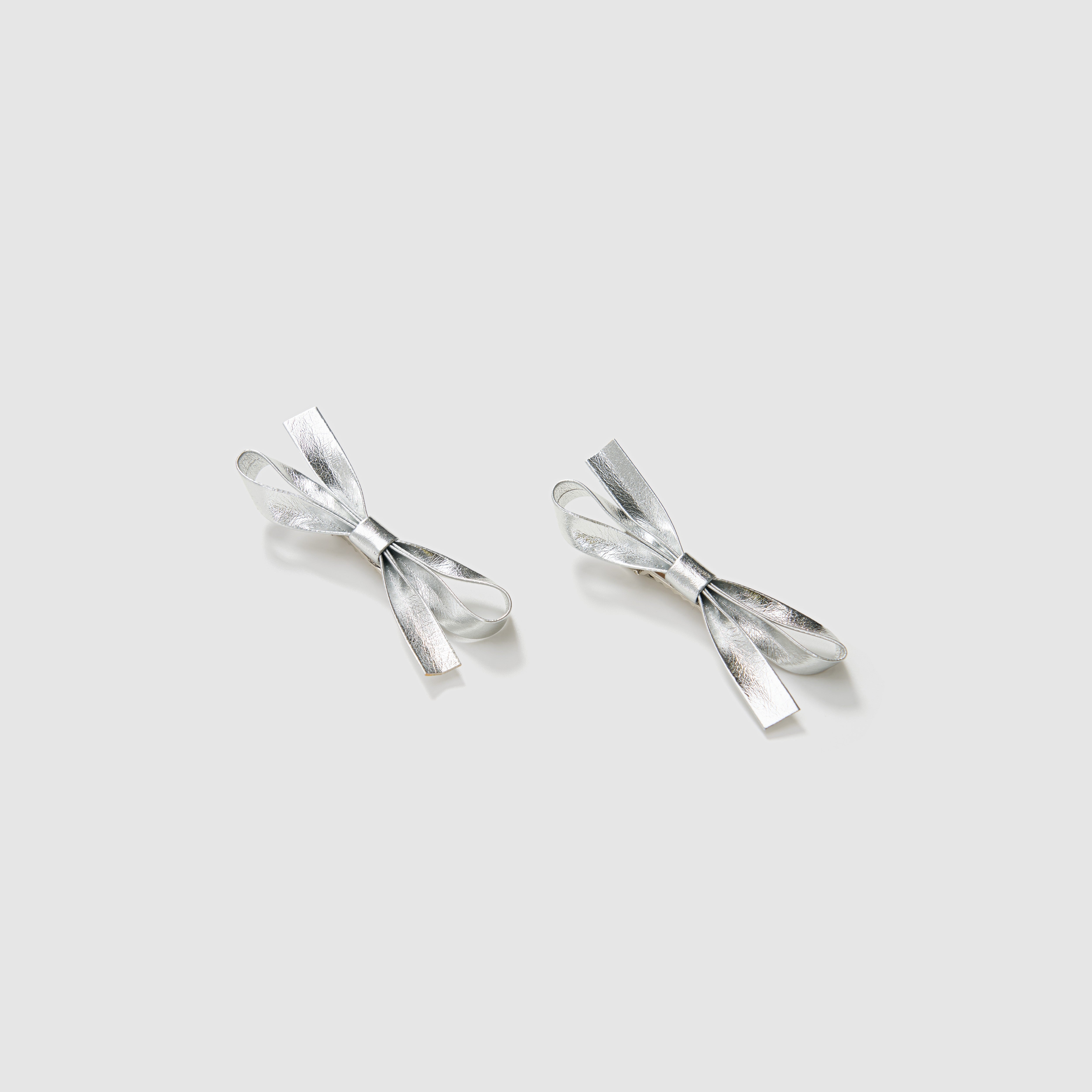 The KIKI and JOJO Girls Silver Bow Hair Clips - Set of 2 feature metallic silver PU leather bows, beautifully displayed on a light gray background—ideal as elegant hair accessories.