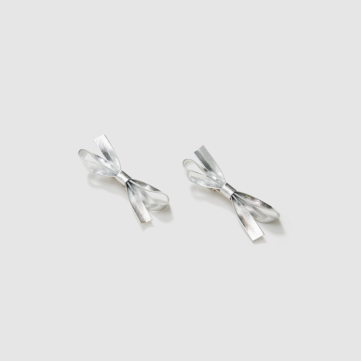 The KIKI and JOJO Girls Silver Bow Hair Clips - Set of 2 feature metallic silver PU leather bows, beautifully displayed on a light gray background—ideal as elegant hair accessories.