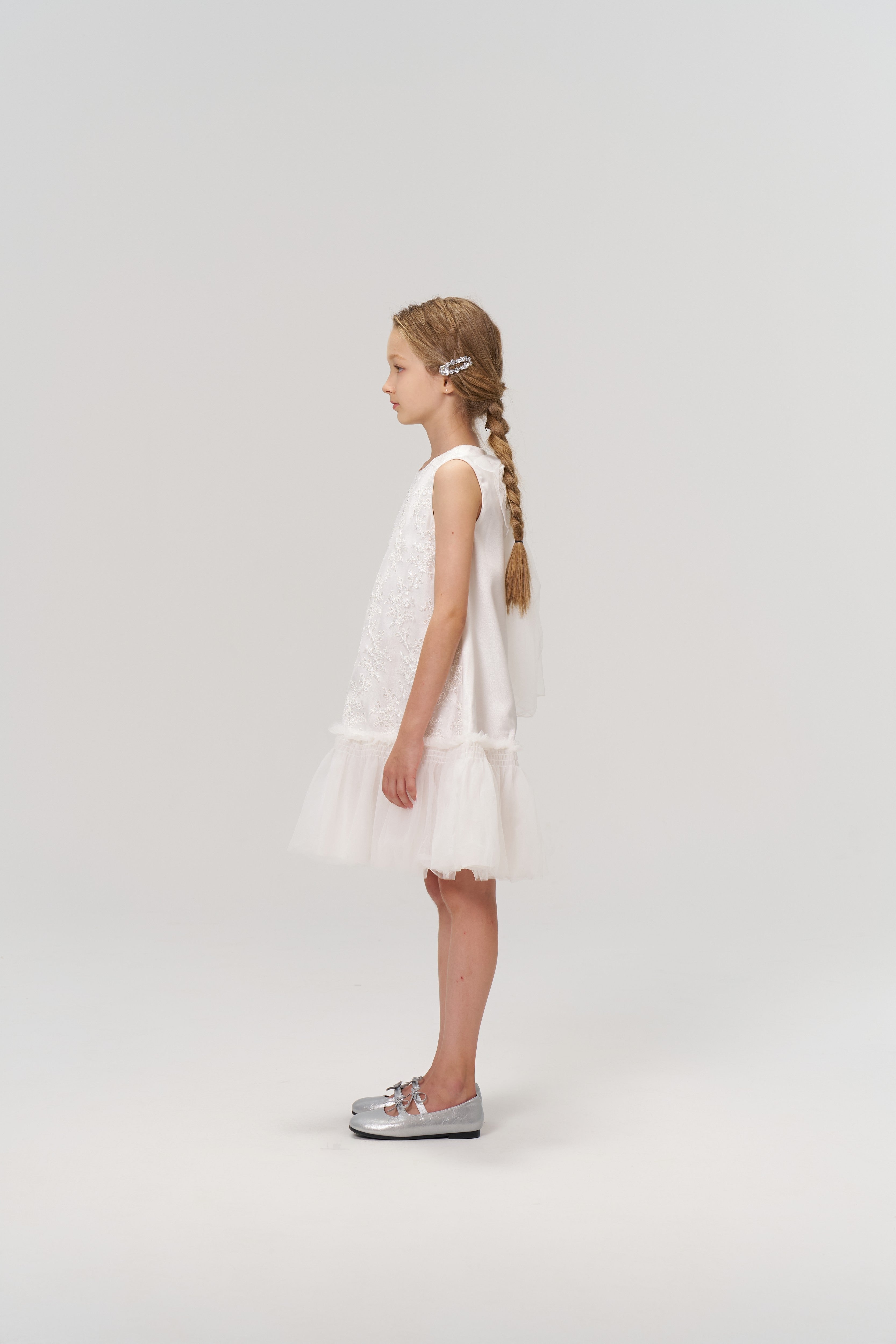 A young girl with long braided hair, styled with KIKI and JOJO’s Girls Crystal Garden Barrettes (set of 2), wears a sleeveless white dress and silver shoes, standing in profile against a plain light background.