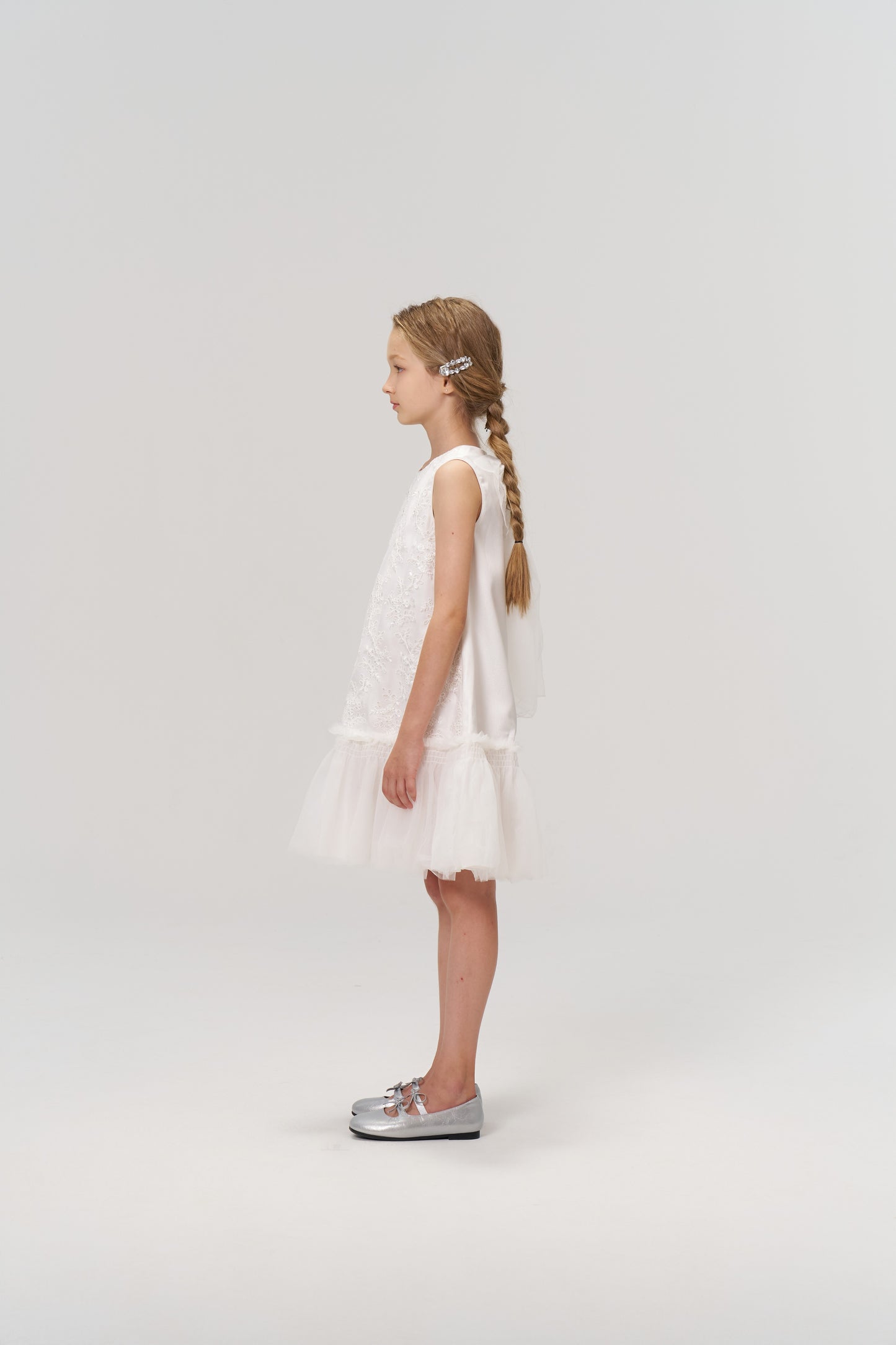 A young girl with long braided hair, styled with KIKI and JOJO’s Girls Crystal Garden Barrettes (set of 2), wears a sleeveless white dress and silver shoes, standing in profile against a plain light background.