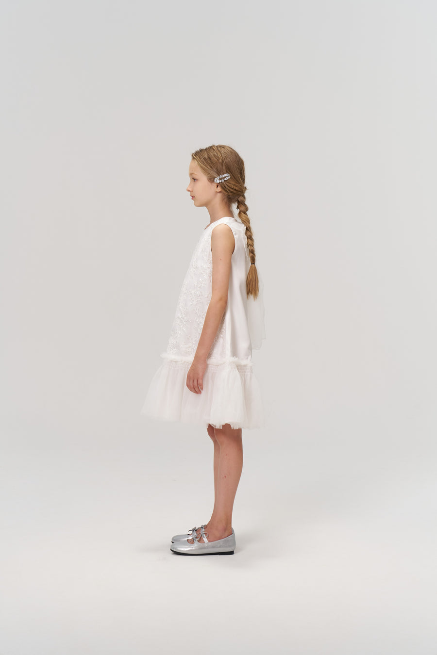 A young girl with long braided hair, styled with KIKI and JOJO’s Girls Crystal Garden Barrettes (set of 2), wears a sleeveless white dress and silver shoes, standing in profile against a plain light background.