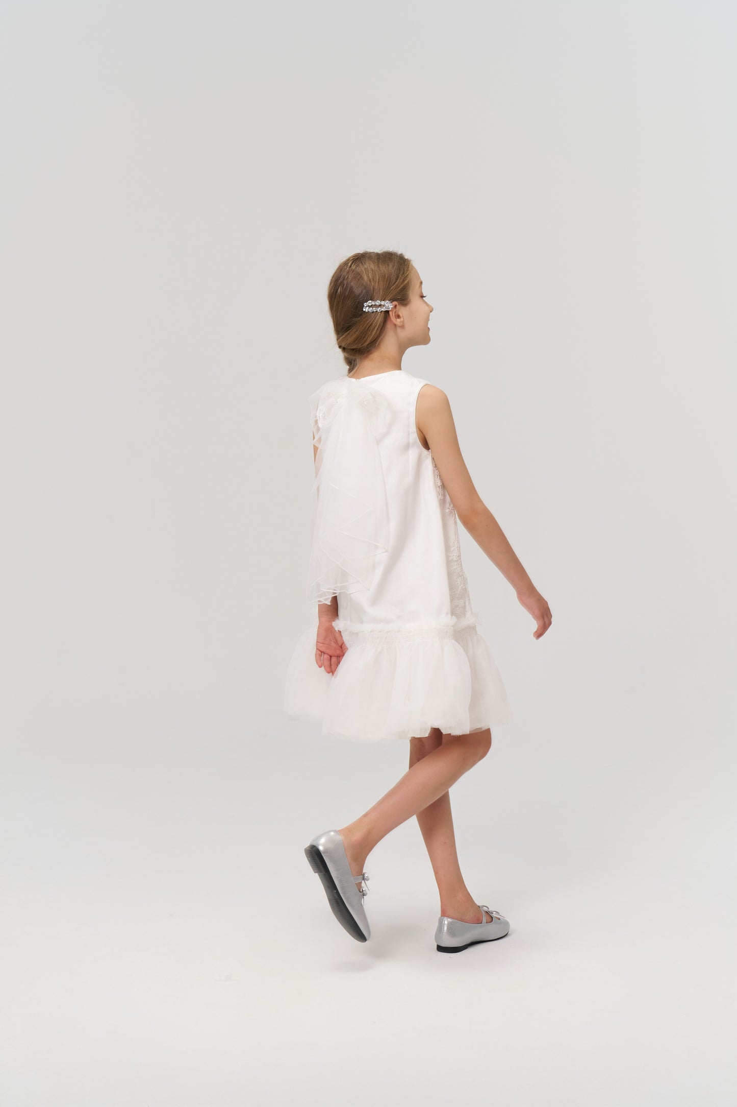 A young girl wearing KIKI and JOJO Girls Crystal Garden Barrettes (set of 2) in her hair, a white dress, and silver shoes walks to the right against a plain white background.