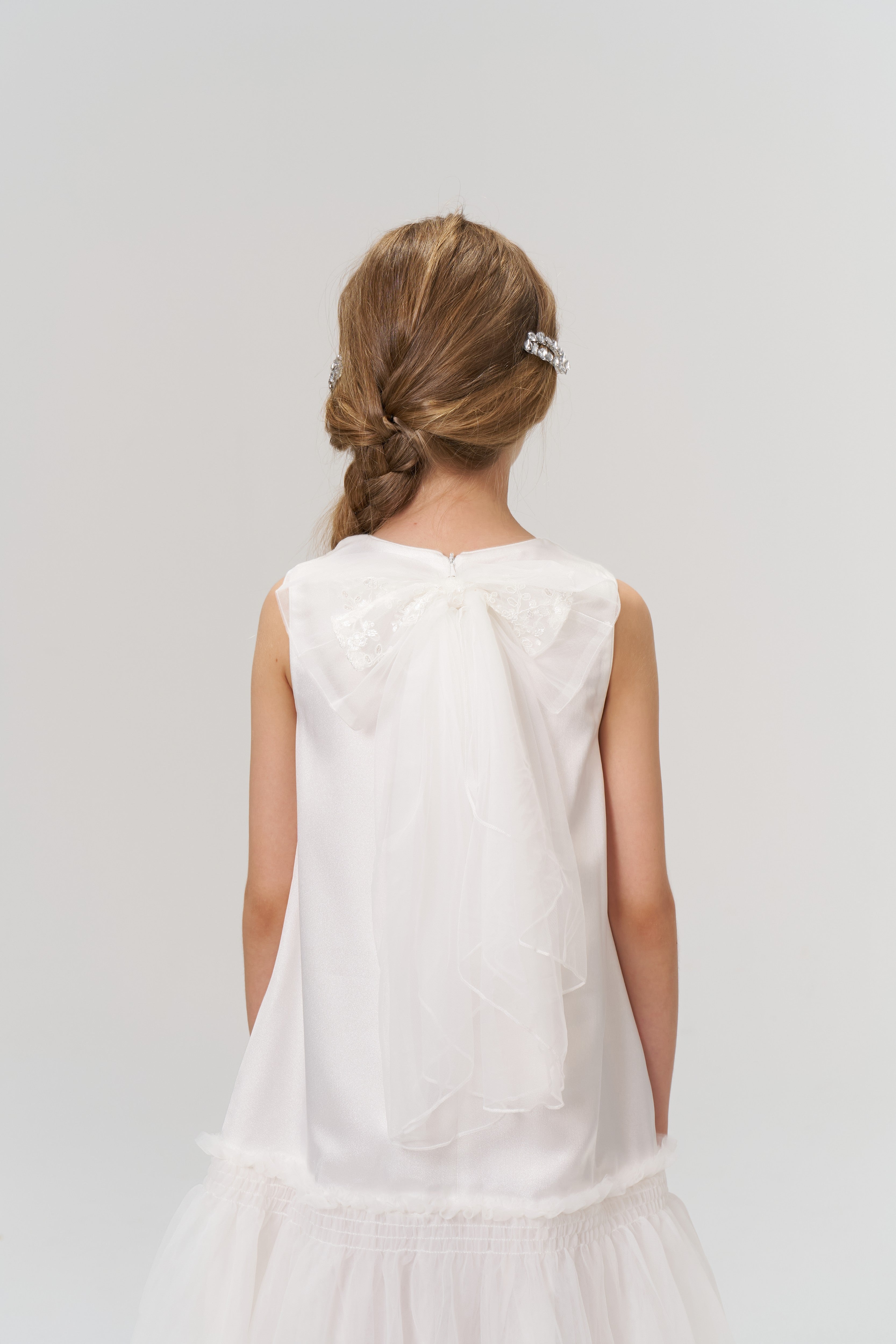 A young girl with light brown hair in a loose braid, styled with KIKI and JOJO Girls Crystal Garden Barrettes (Set of 2), is shown from behind wearing a white dress with a sheer bow and layered tulle skirt.