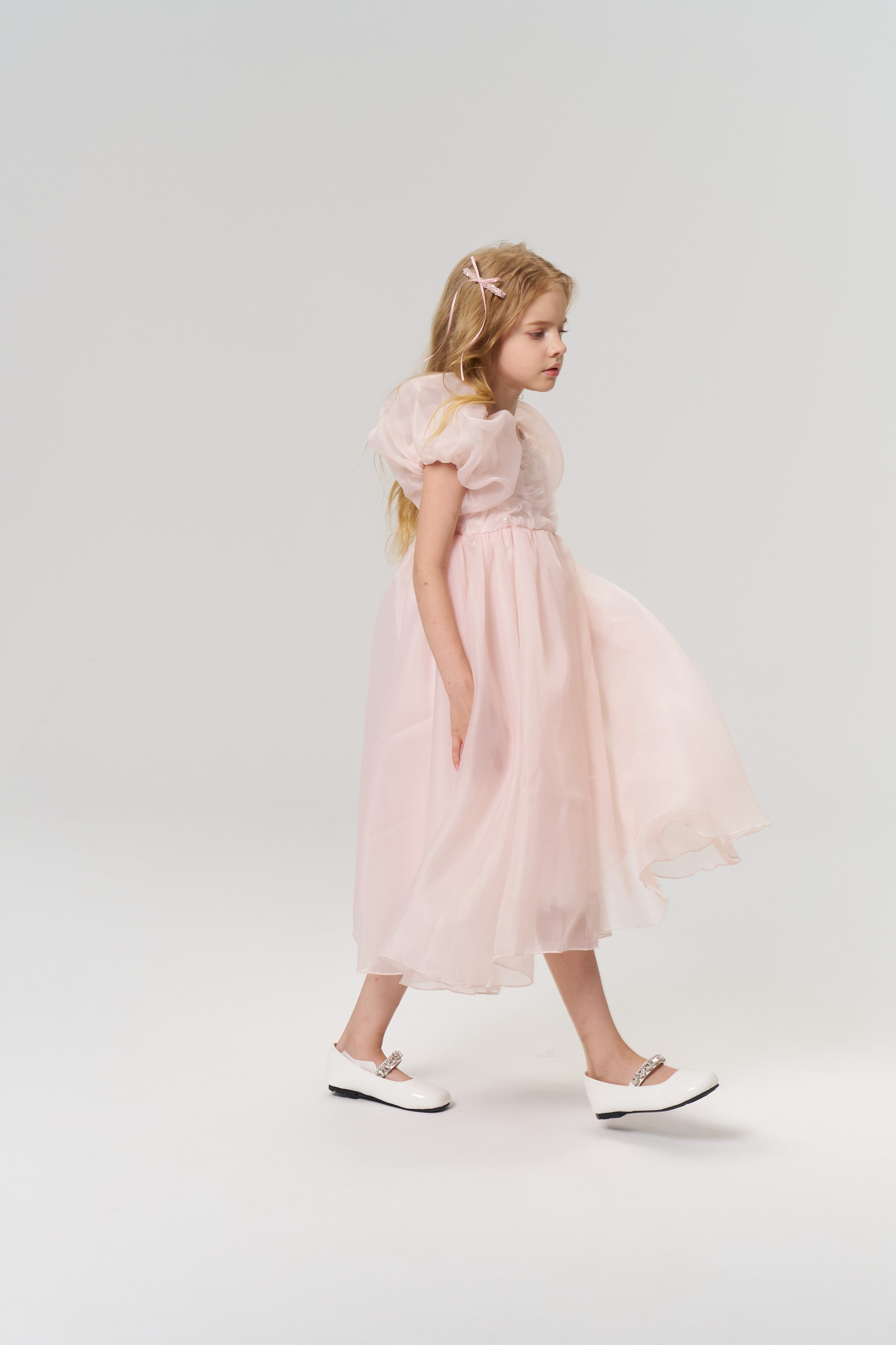 A young girl with long blonde hair, accessorized with KIKI and JOJO's Girls Pink Beaded Bow Hair Clips (Set of 2), walks left in a light pink dress and white shoes against a plain light background.
