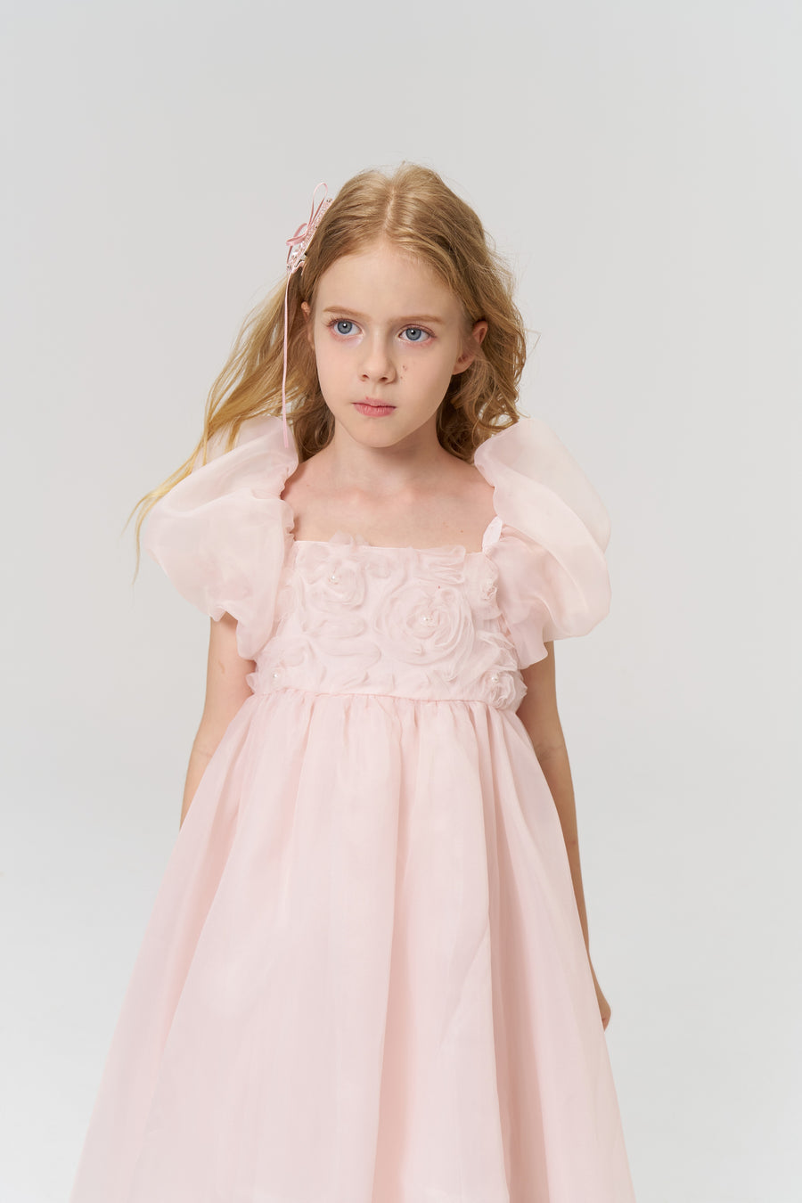 A young girl with long blonde hair styled with KIKI and JOJO’s Girls Pink Beaded Bow Hair Clips (Set of 2) wears a light pink floral dress, standing against a plain light background.