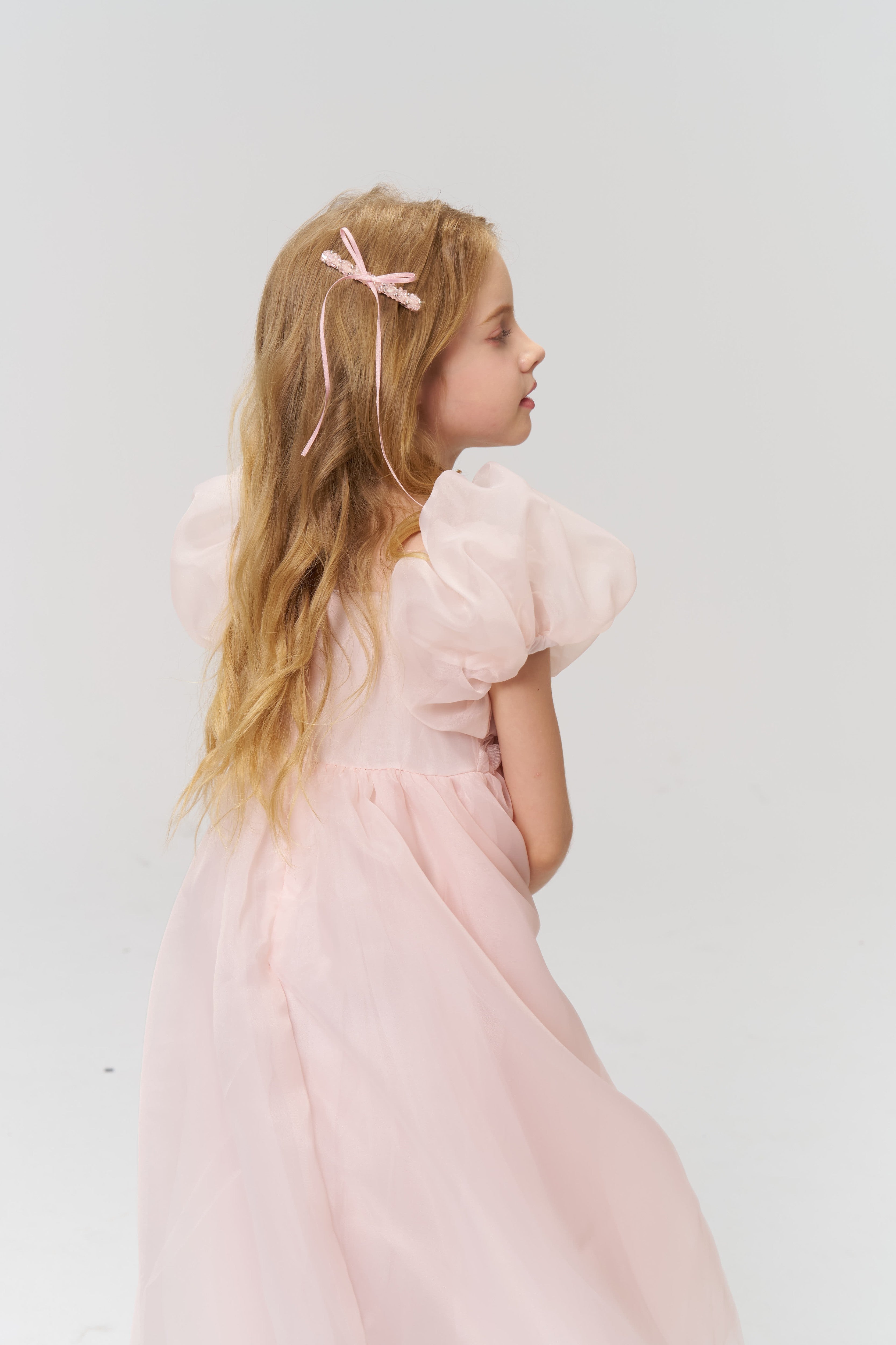 A young girl with long blonde hair wears a light pink dress and KIKI and JOJO’s Girls Pink Beaded Bow Hair Clip from the set of 2, standing sideways against a plain light background.