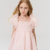 Young girl with long blonde hair styled with KIKI and JOJO Girls Black Beaded Bow Hair Clips (Set of 2), wearing a light pink dress, stands against a plain white background.