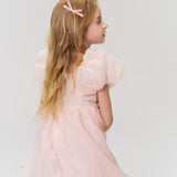 A young girl with long blonde hair, wearing a light pink dress and a black beaded bow hair clip from the Girls Black Beaded Bow Hair Clips - Set of 2 by KIKI and JOJO, stands slightly turned against a plain light background.