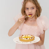 A young girl in a light pink dress wears a KIKI and JOJO Girls Black Beaded Bow Hair Clip, holding a plate with tart and eating dessert against a plain white background.