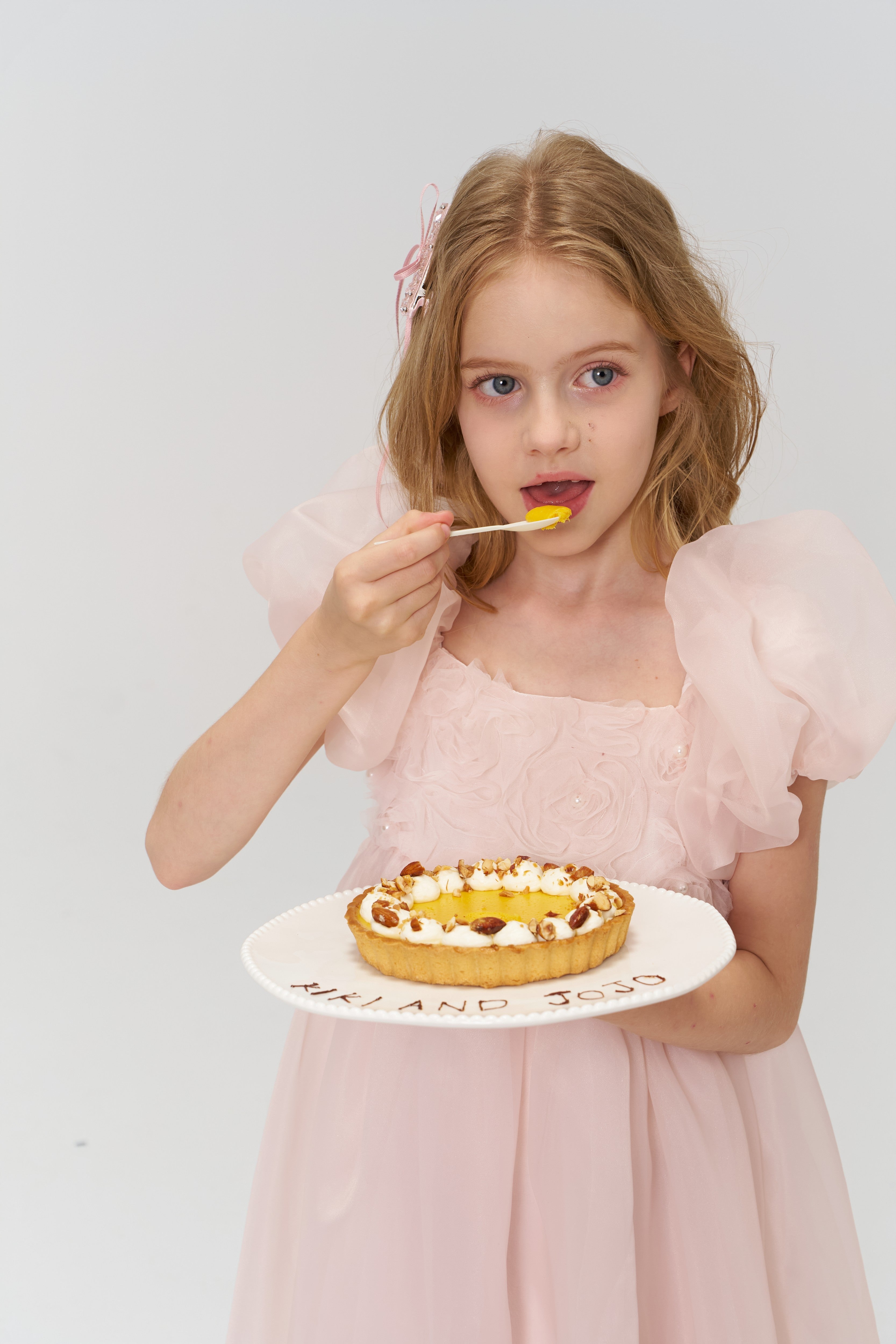 A young girl in a light pink dress wears a KIKI and JOJO Girls Black Beaded Bow Hair Clip, holding a plate with tart and eating dessert against a plain white background.
