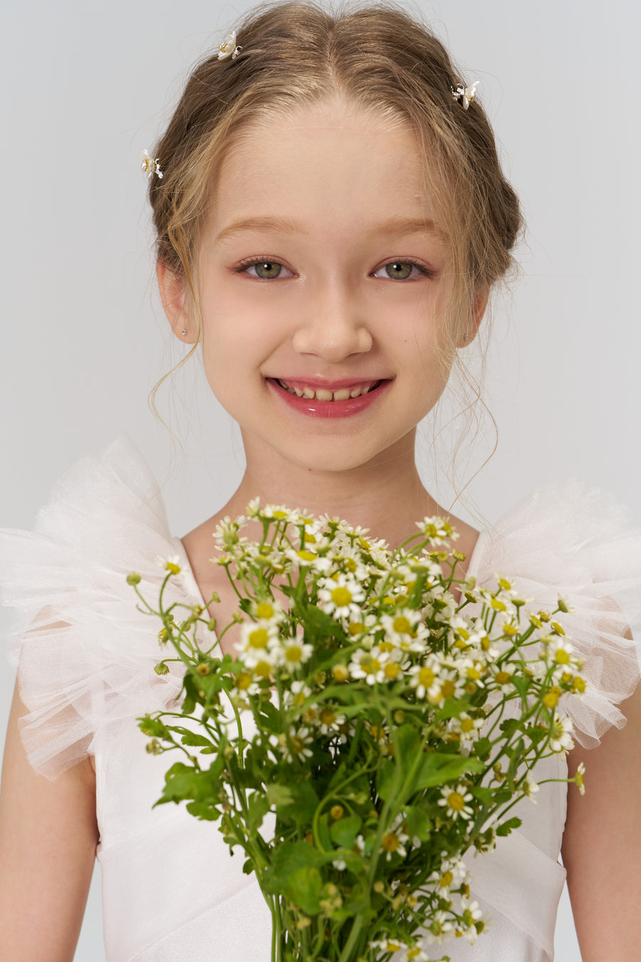 A young blonde girl in a white dress smiles at the camera, holding a bouquet and wearing KIKI and JOJO's Girls Daisy Crystal Flower Hair Clips (Set of 6)—the perfect flower girl hair accessories—against a plain background.