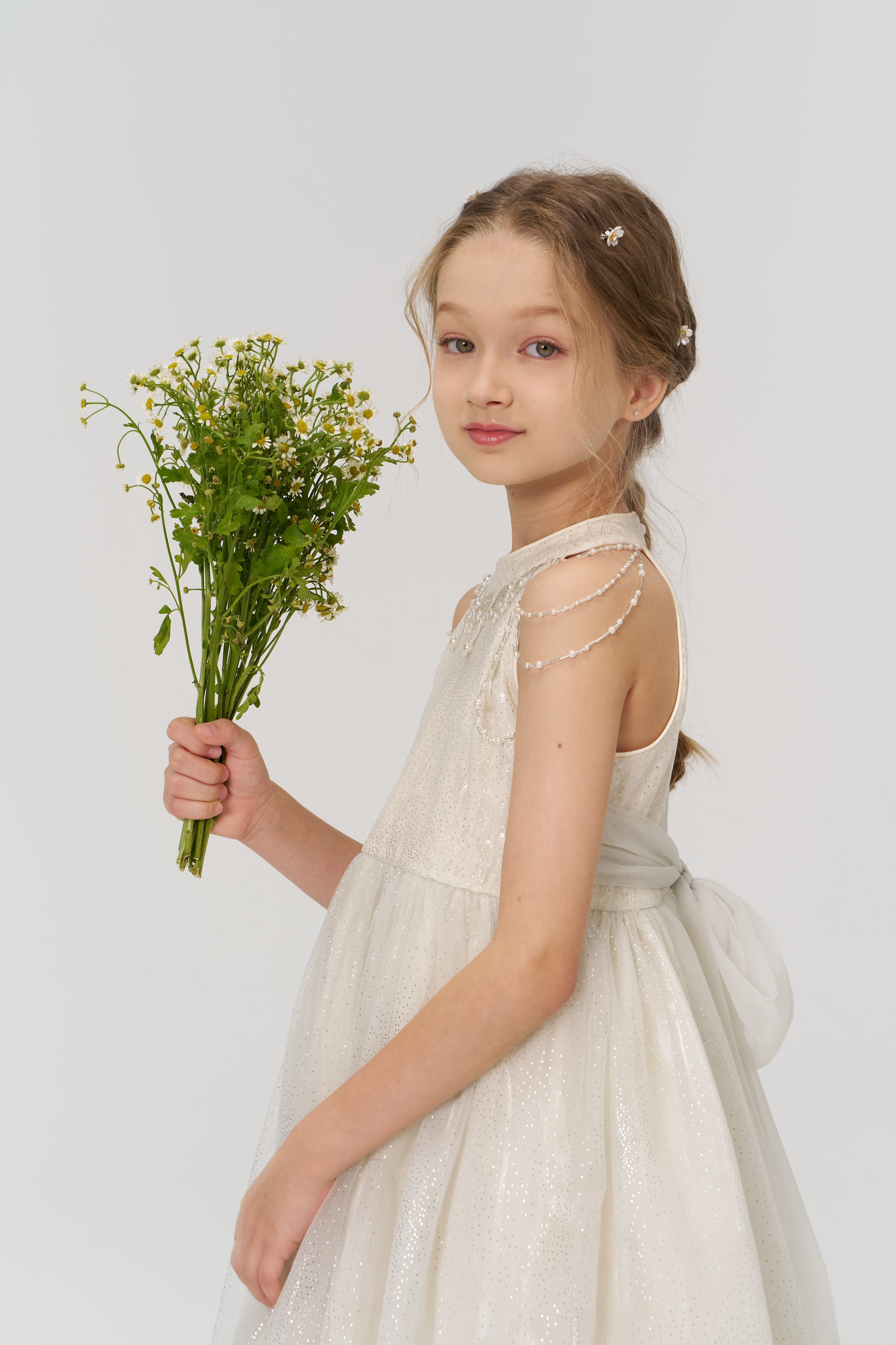 A young girl wears KIKI and JOJO's Girls Daisy Crystal Flower Hair Clips (Set of 6) in her hair, paired with a white dress and a bouquet of yellow and white flowers, standing against a plain light background.