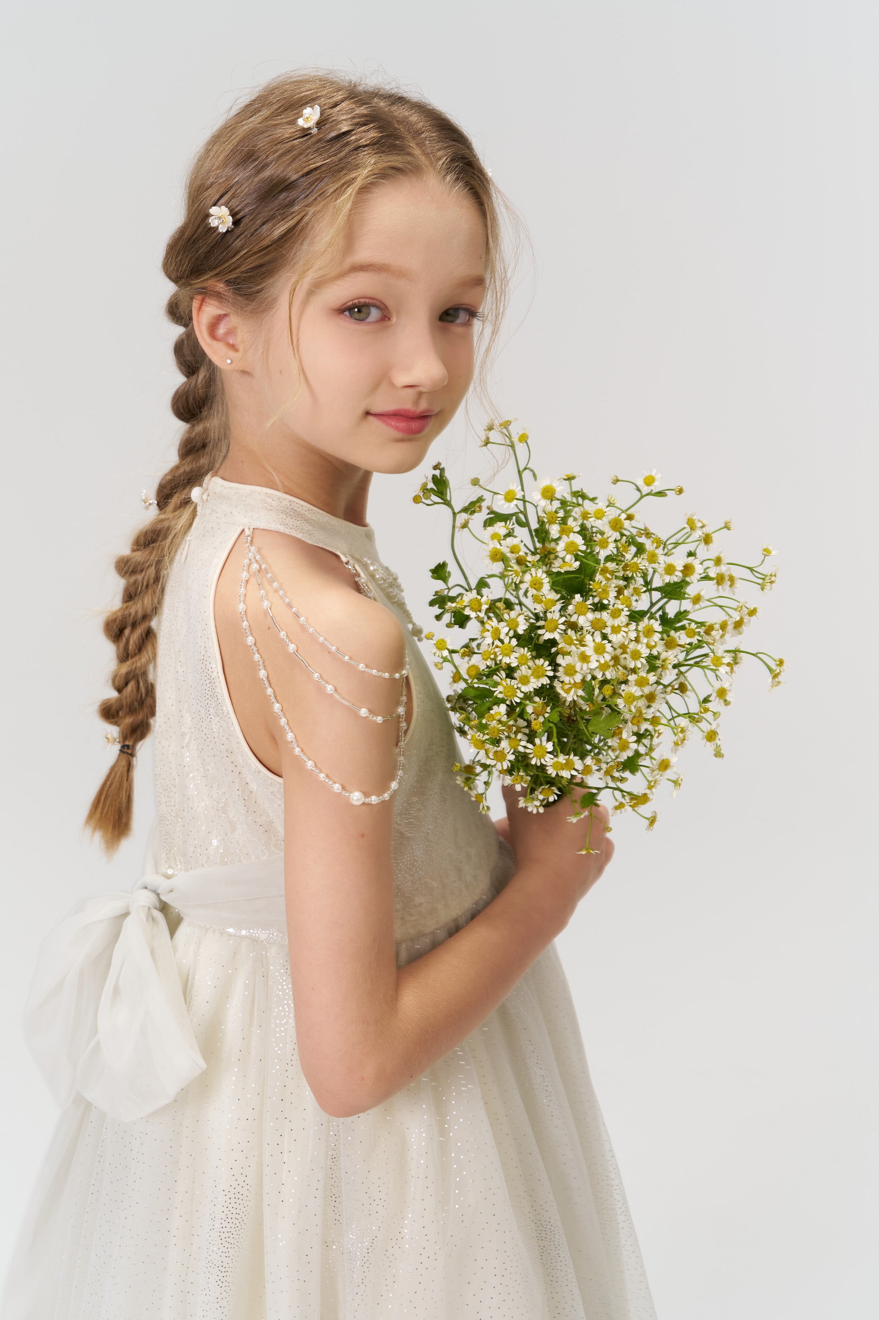 A young girl in a white dress, her braids styled with KIKI and JOJO’s Girls Daisy Crystal Flower Hair Clips (Set of 6), holds a bouquet of small white and yellow flowers against a plain light background.