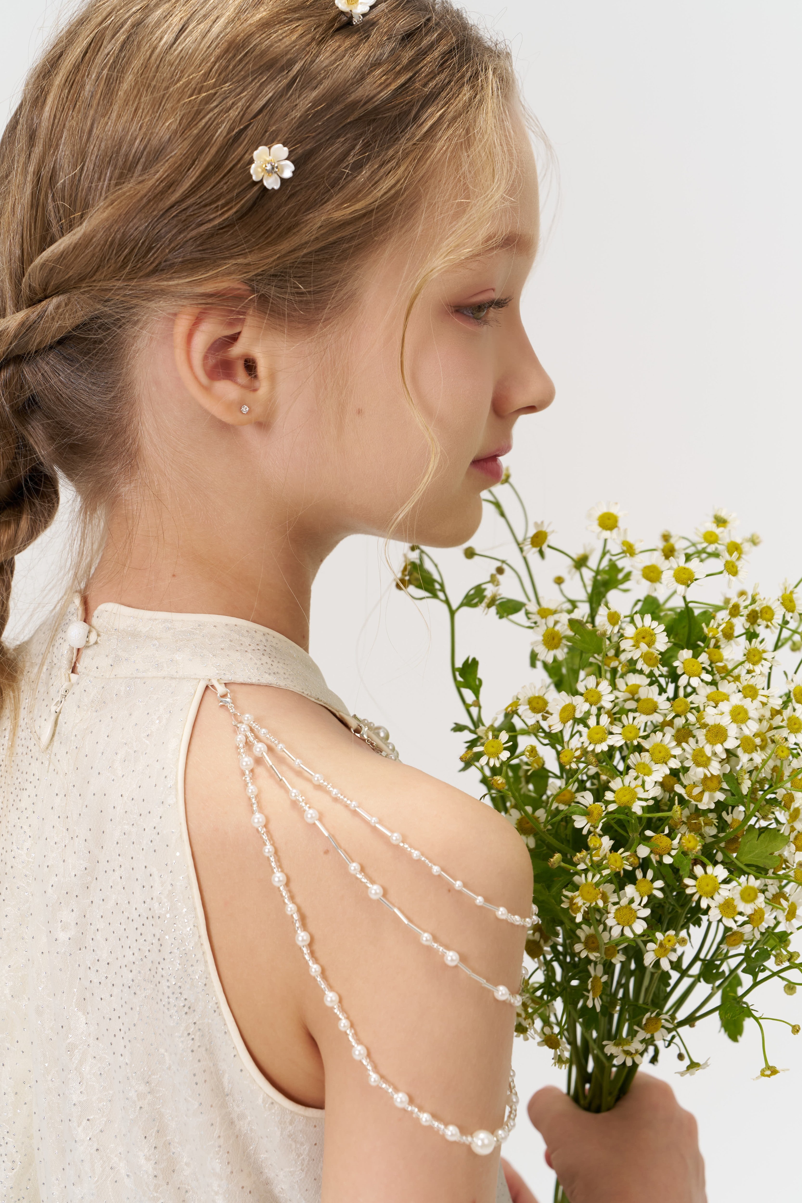 A young girl with braided hair styled with KIKI and JOJO’s Girls Daisy Crystal Flower Hair Clips (set of 6) wears a cream dress and beaded shoulder accessory, holding chamomile flowers as she looks to the side.