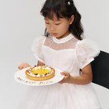 A young girl in a white dress sits on a black chair, holding a "HAPPY ANN JOJO" cake, her hair decorated with KIKI and JOJO Girls Daisy Crystal Flower Hair Clips – Set of 6.