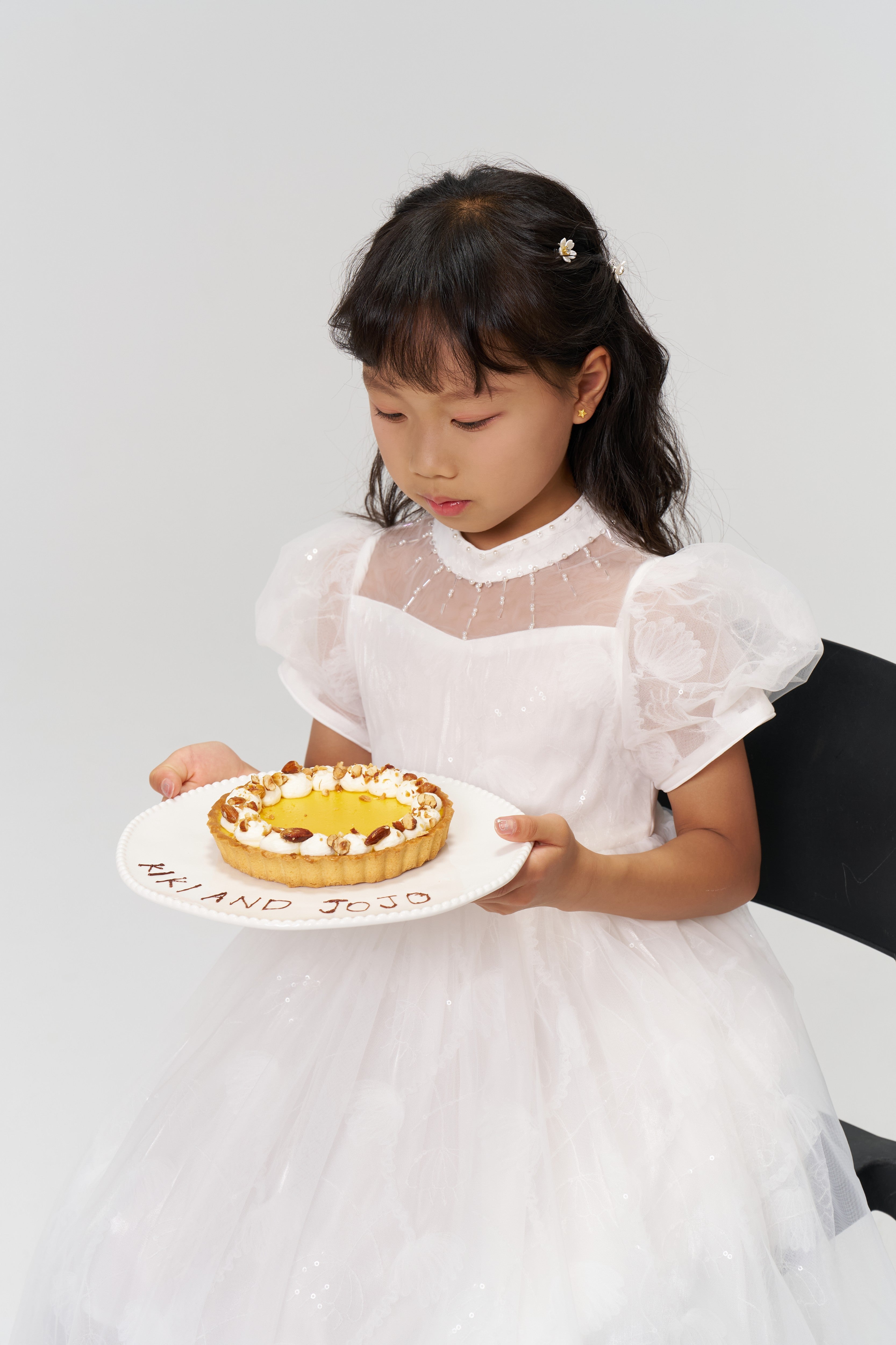 A young girl in a white dress sits on a black chair, holding a "HAPPY ANN JOJO" cake, her hair decorated with KIKI and JOJO Girls Daisy Crystal Flower Hair Clips – Set of 6.