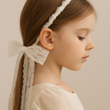 A young girl in a cream dress wears the KIKI and JOJO Girls Pearl Lace Ribbon Headband, featuring a large white lace bow, against a neutral background.