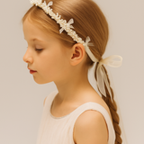 A young girl in a light sleeveless dress wears the KIKI and JOJO Girls Pearl Bow Crown Headband, featuring organza bows and pearls, her long hair braided and shown in profile against a plain background.