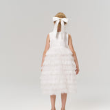 A young flower girl faces away in a white dress and shoes, her hair adorned with the KIKI and JOJO Girls Hand-Beaded Pearl Satin Bow Hair Clip, set against a plain light background.