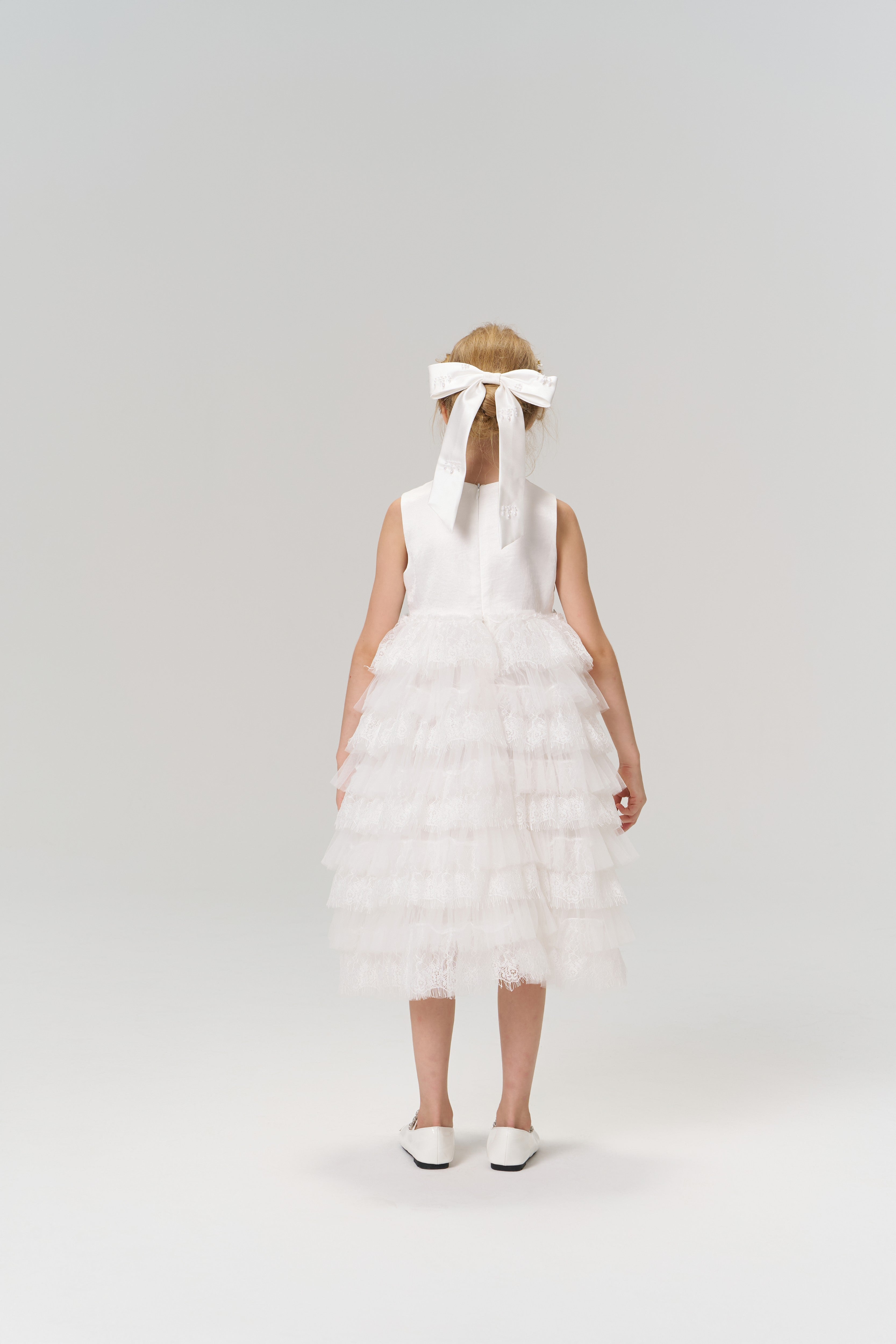 A young flower girl faces away in a white dress and shoes, her hair adorned with the KIKI and JOJO Girls Hand-Beaded Pearl Satin Bow Hair Clip, set against a plain light background.