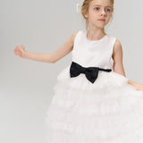 A young flower girl wearing the Girls Hand-Beaded Pearl Satin Bow Hair Clip by KIKI and JOJO stands with one arm outstretched against a plain, light background.