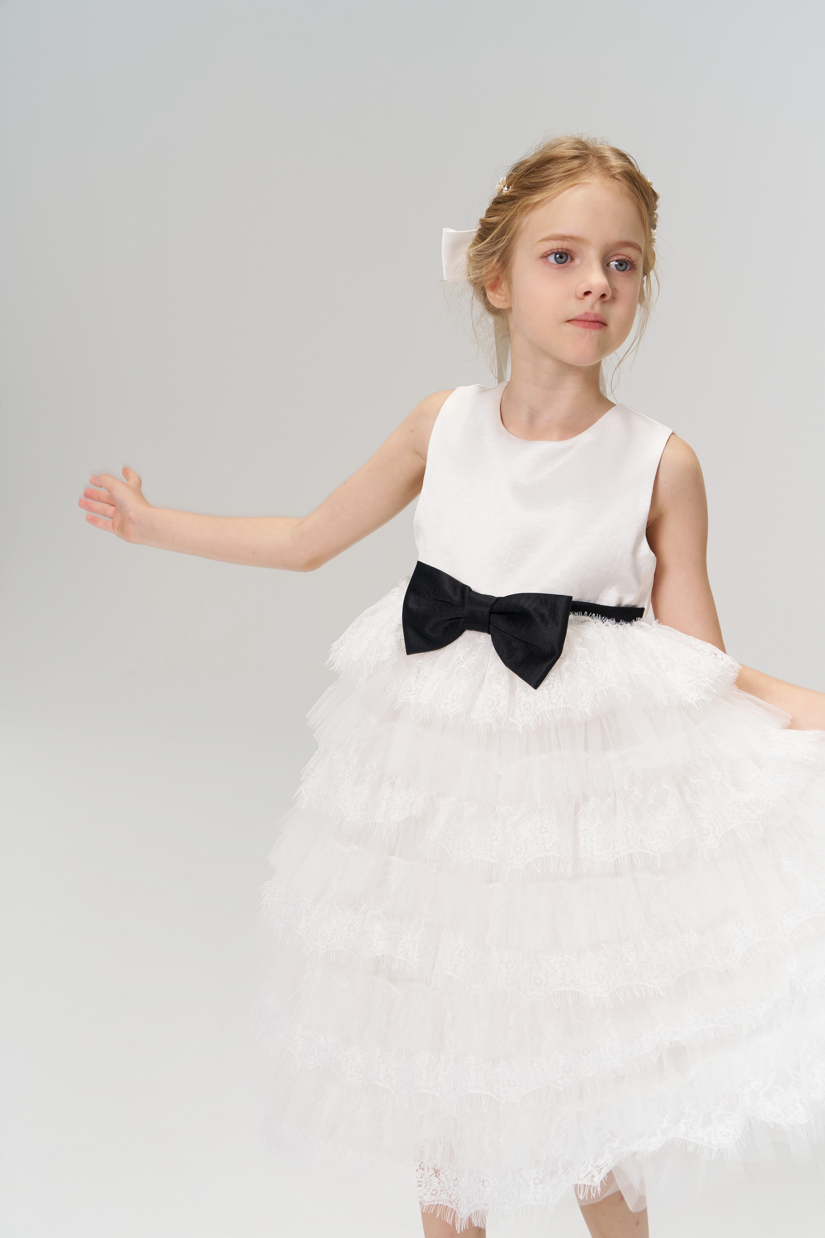 A young flower girl wearing the Girls Hand-Beaded Pearl Satin Bow Hair Clip by KIKI and JOJO stands with one arm outstretched against a plain, light background.