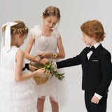 Three children stand together: two flower girls in white, one wearing the KIKI and JOJO Girls Hand-Beaded Pearl Satin Bow Hair Clip, hold a flower basket, while a boy in a black suit reaches toward it.