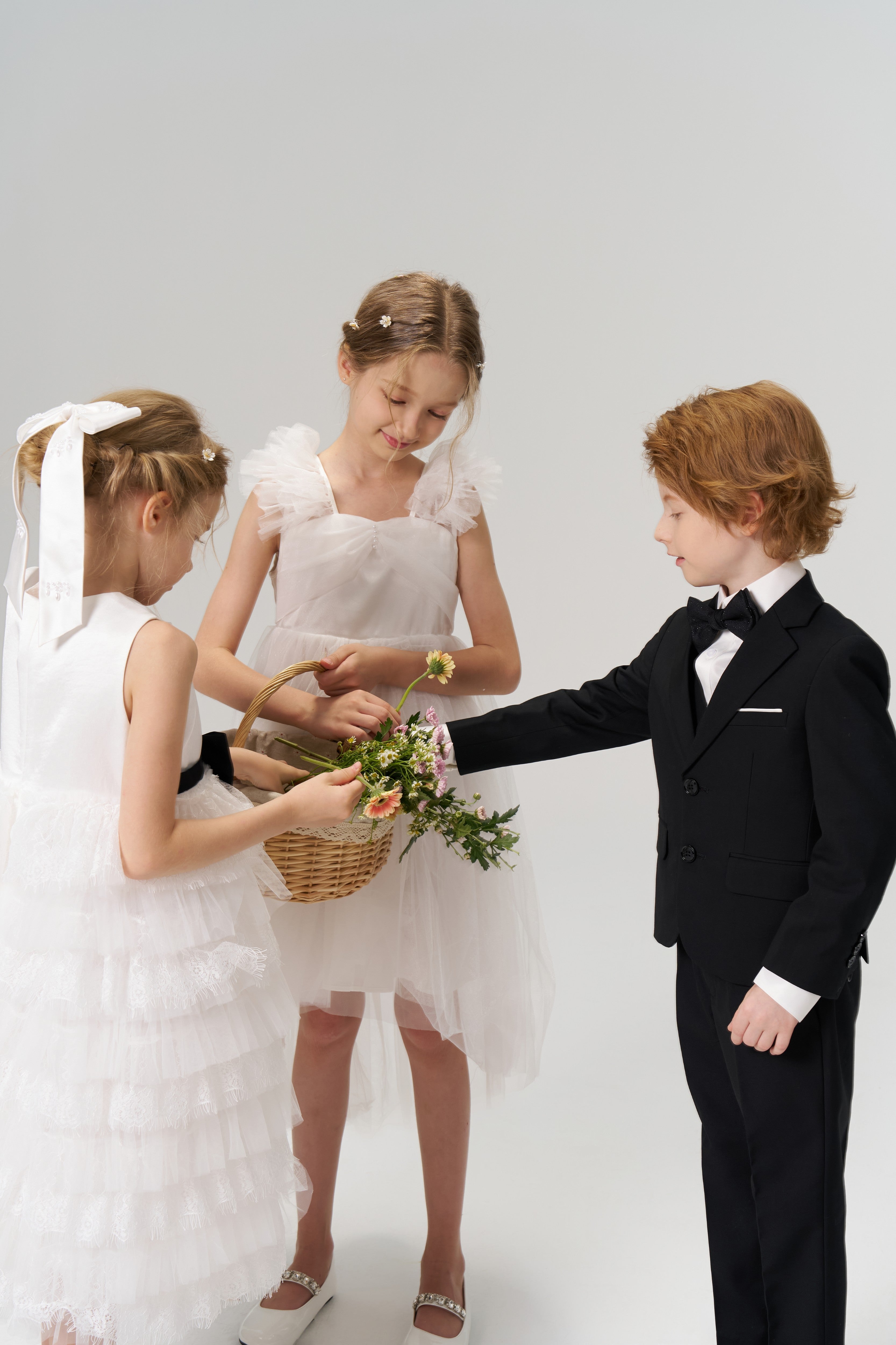 Three children stand together: two flower girls in white, one wearing the KIKI and JOJO Girls Hand-Beaded Pearl Satin Bow Hair Clip, hold a flower basket, while a boy in a black suit reaches toward it.