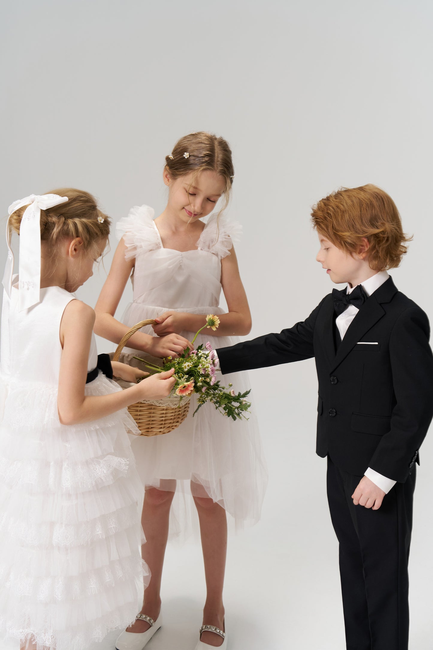 Three children stand together: two flower girls in white, one wearing the KIKI and JOJO Girls Hand-Beaded Pearl Satin Bow Hair Clip, hold a flower basket, while a boy in a black suit reaches toward it.