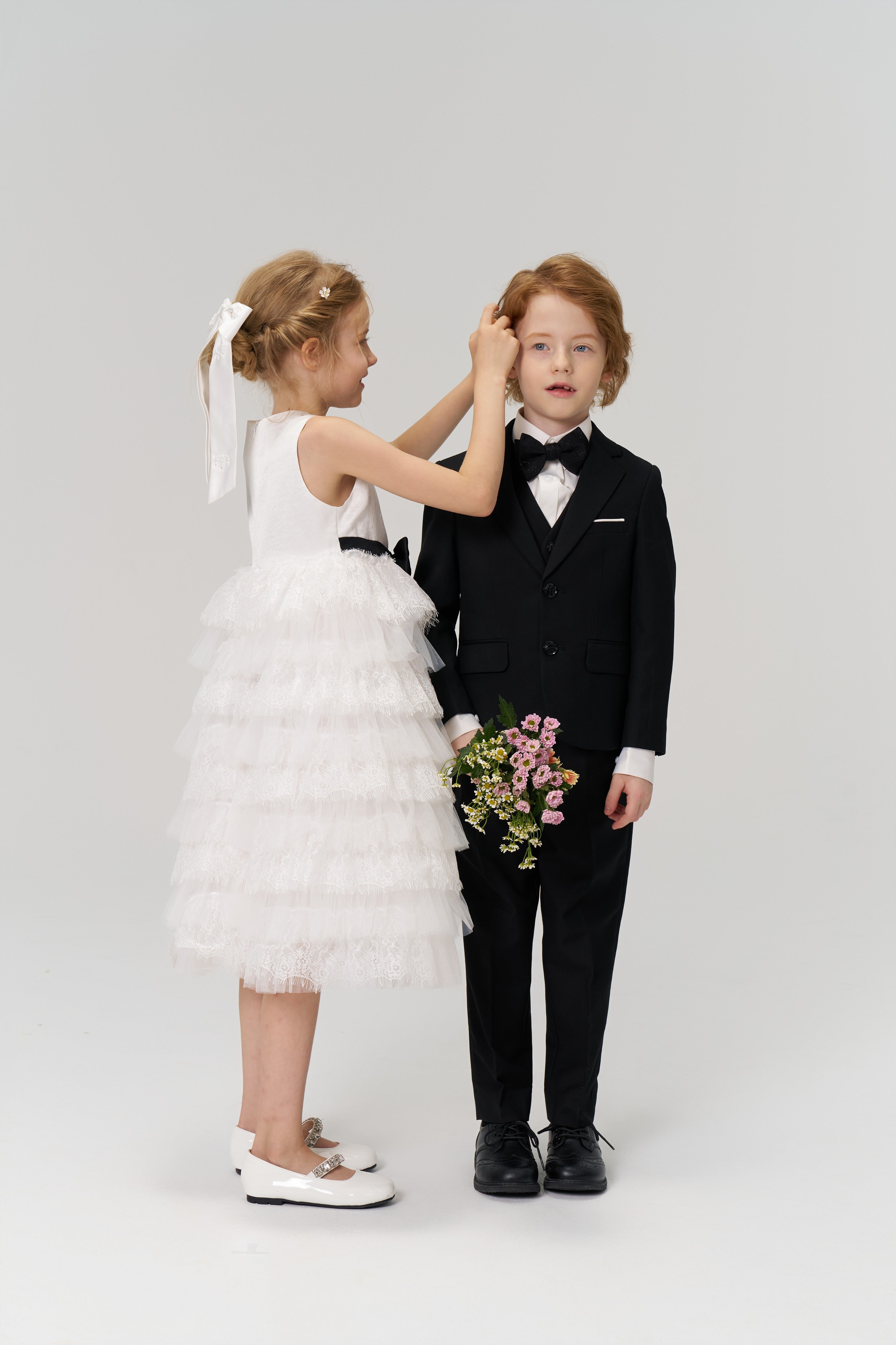 A girl wearing the KIKI and JOJO Girls Hand-Beaded Pearl Satin Bow Hair Clip adjusts a boy’s hair as he holds a bouquet; both are dressed formally against a plain background.