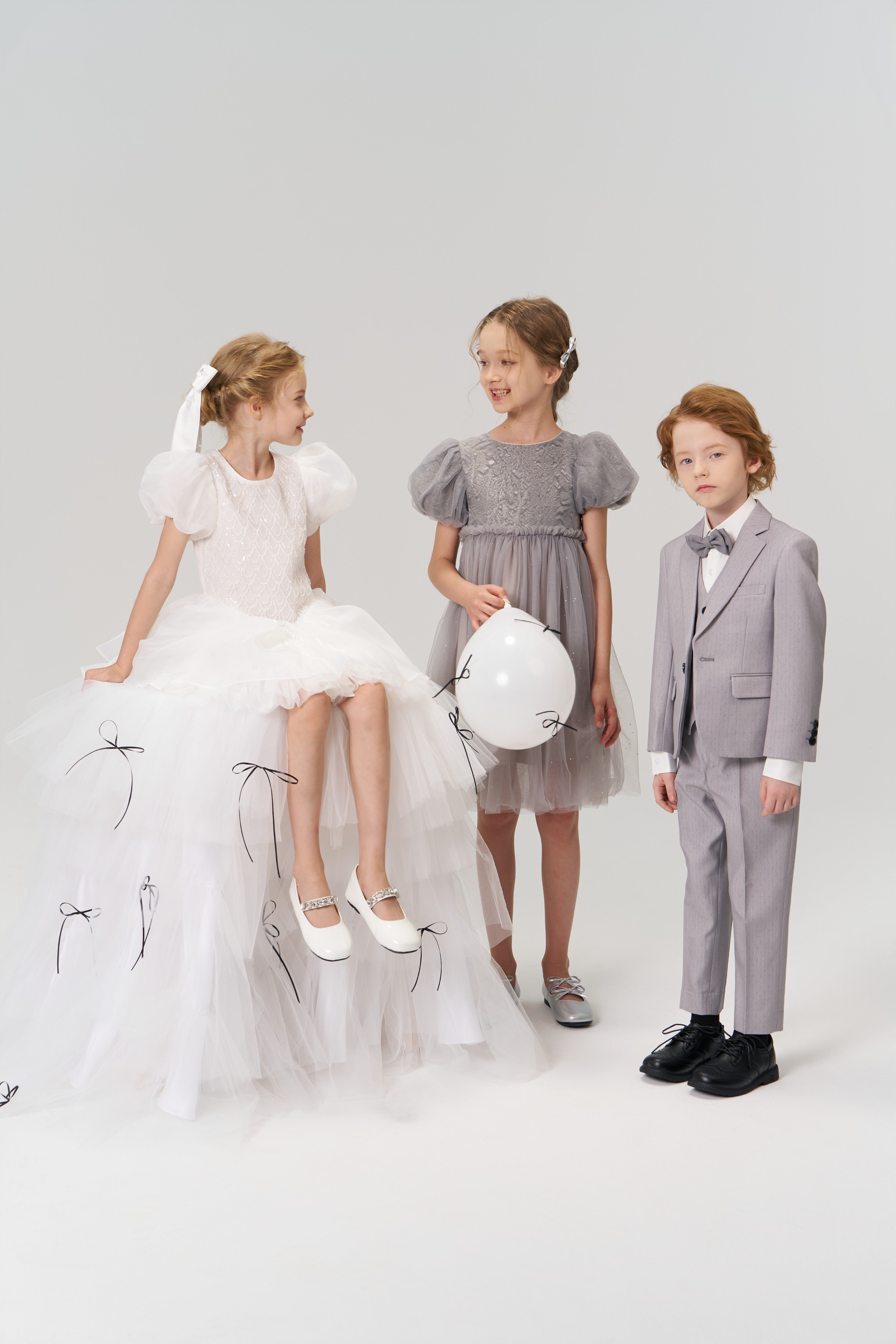 Three children pose formally: a flower girl with a white balloon wears KIKI and JOJO's Girls Hand-Beaded Pearl Satin Bow Hair Clip, another girl sits nearby, and a boy in a suit stands beside them against a plain background.