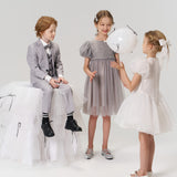 Three children dressed formally, including a flower girl wearing the KIKI and JOJO Girls Hand-Beaded Pearl Satin Bow Hair Clip, play with a white balloon in a minimalist studio setting.