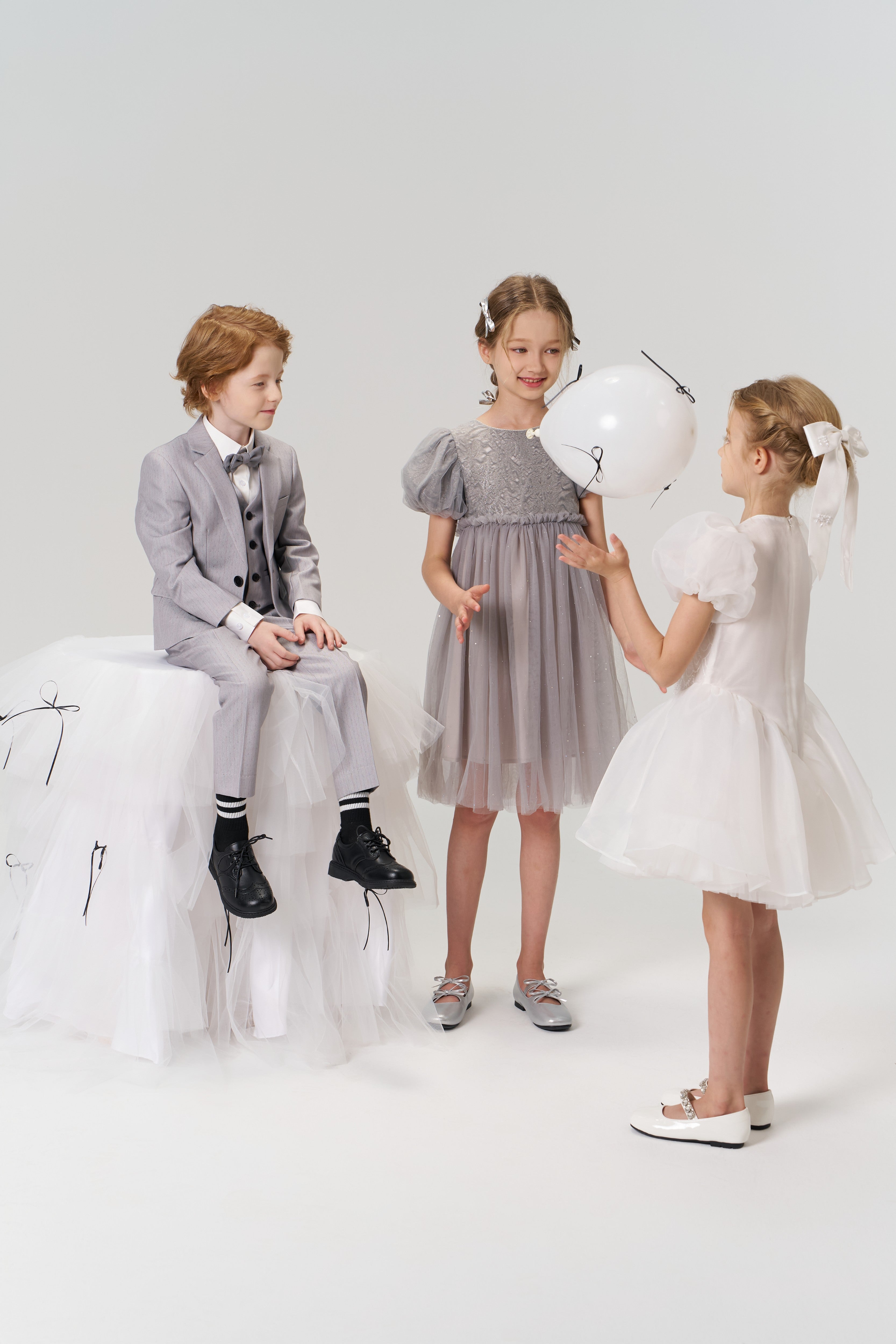 Three children dressed formally, including a flower girl wearing the KIKI and JOJO Girls Hand-Beaded Pearl Satin Bow Hair Clip, play with a white balloon in a minimalist studio setting.