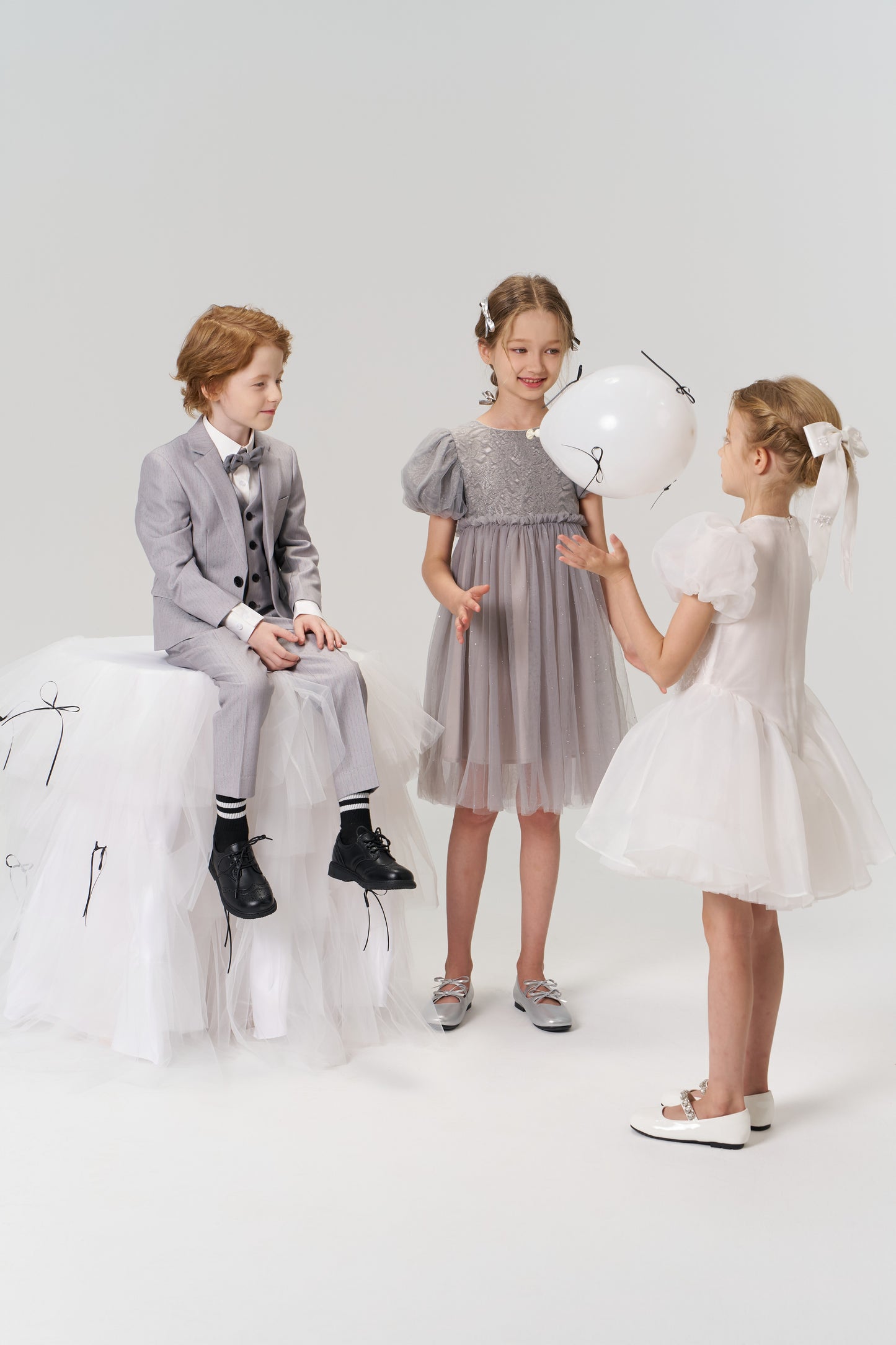 Three children dressed formally, including a flower girl wearing the KIKI and JOJO Girls Hand-Beaded Pearl Satin Bow Hair Clip, play with a white balloon in a minimalist studio setting.