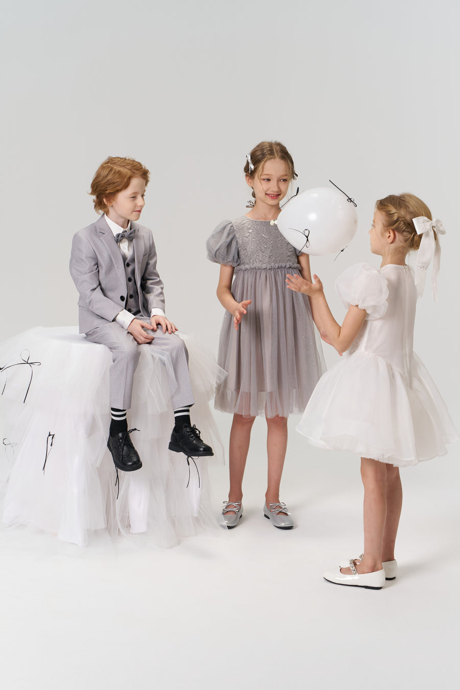 Three children dressed formally, including a flower girl wearing the KIKI and JOJO Girls Hand-Beaded Pearl Satin Bow Hair Clip, play with a white balloon in a minimalist studio setting.