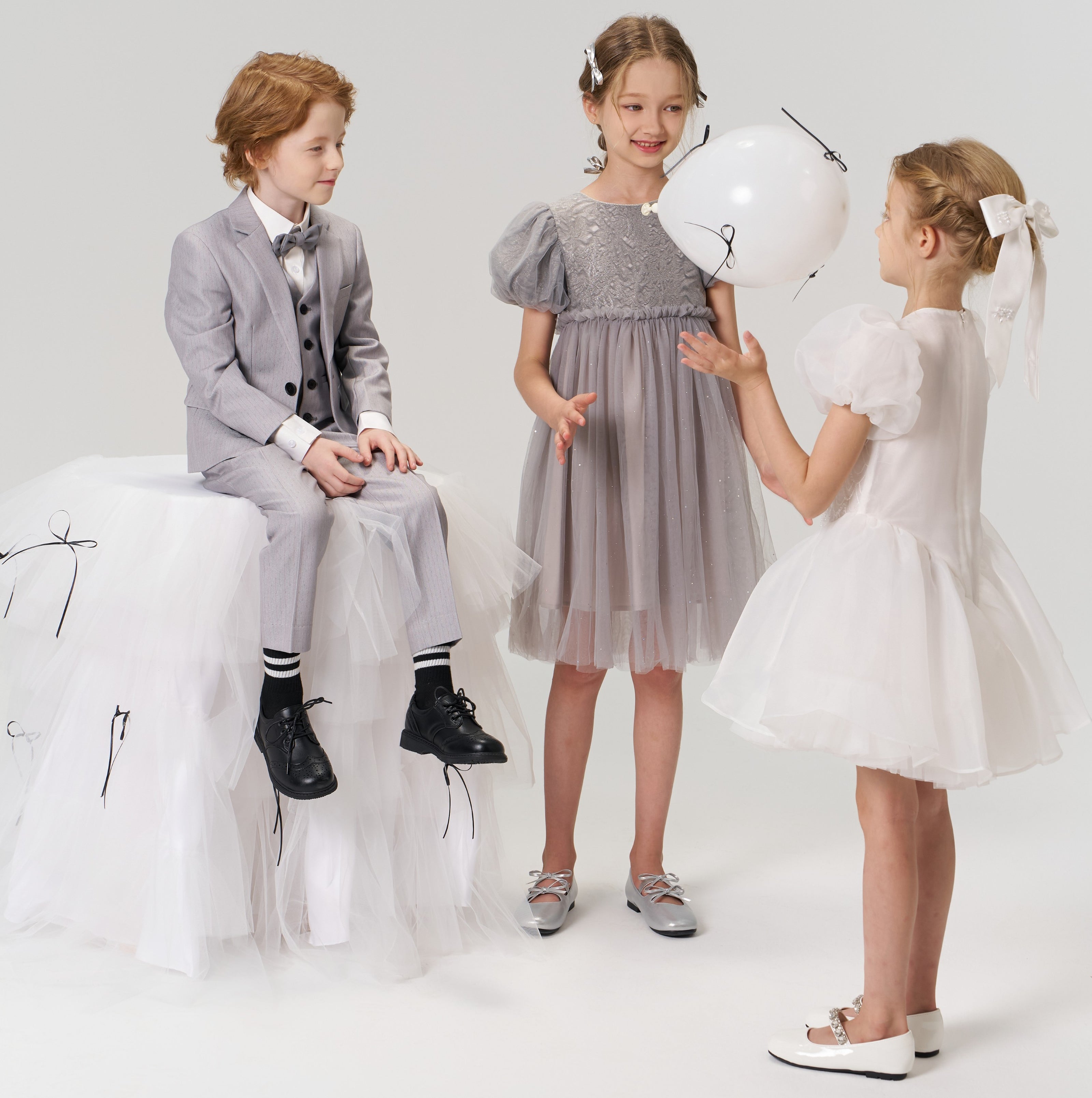 Three children dressed in formal attire stand together; one boy in a suit sits on a white tulle-covered block while two girls in dresses, one holding a white balloon, stand in front of him.
