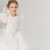 A young flower girl wears a KIKI and JOJO Girls Hand-Beaded Pearl Satin Bow Hair Clip, standing in a white dress with puffed sleeves against a plain, light background.