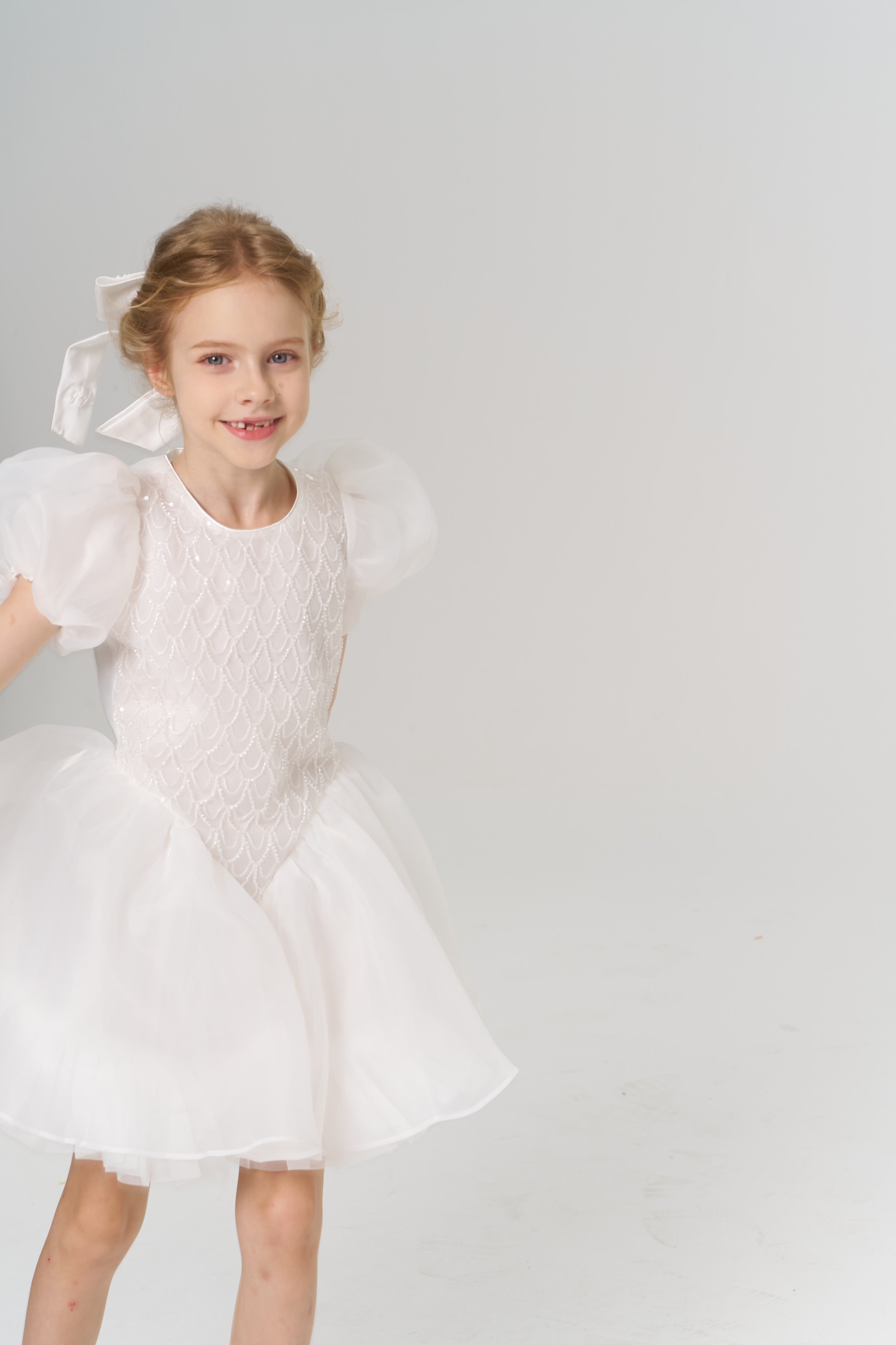 A young flower girl wears a KIKI and JOJO Girls Hand-Beaded Pearl Satin Bow Hair Clip, standing in a white dress with puffed sleeves against a plain, light background.