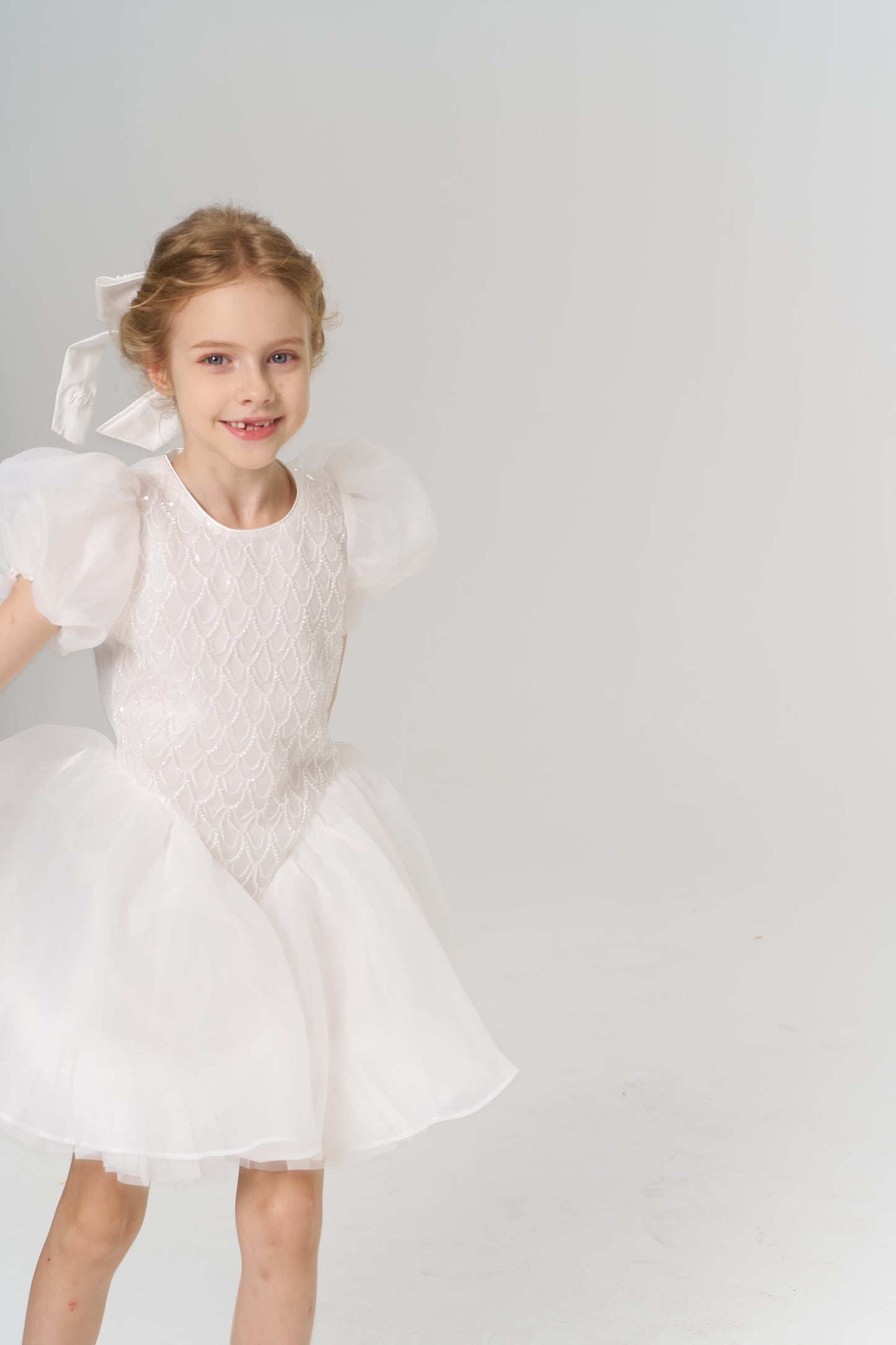 A young flower girl wears a KIKI and JOJO Girls Hand-Beaded Pearl Satin Bow Hair Clip, standing in a white dress with puffed sleeves against a plain, light background.