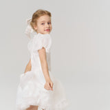 A young girl with a beaming smile wears the KIKI and JOJO Girls Hand-Beaded Pearl Satin Bow Hair Clip, pairing it beautifully with her white puffed-sleeve dress against a simple, light background.