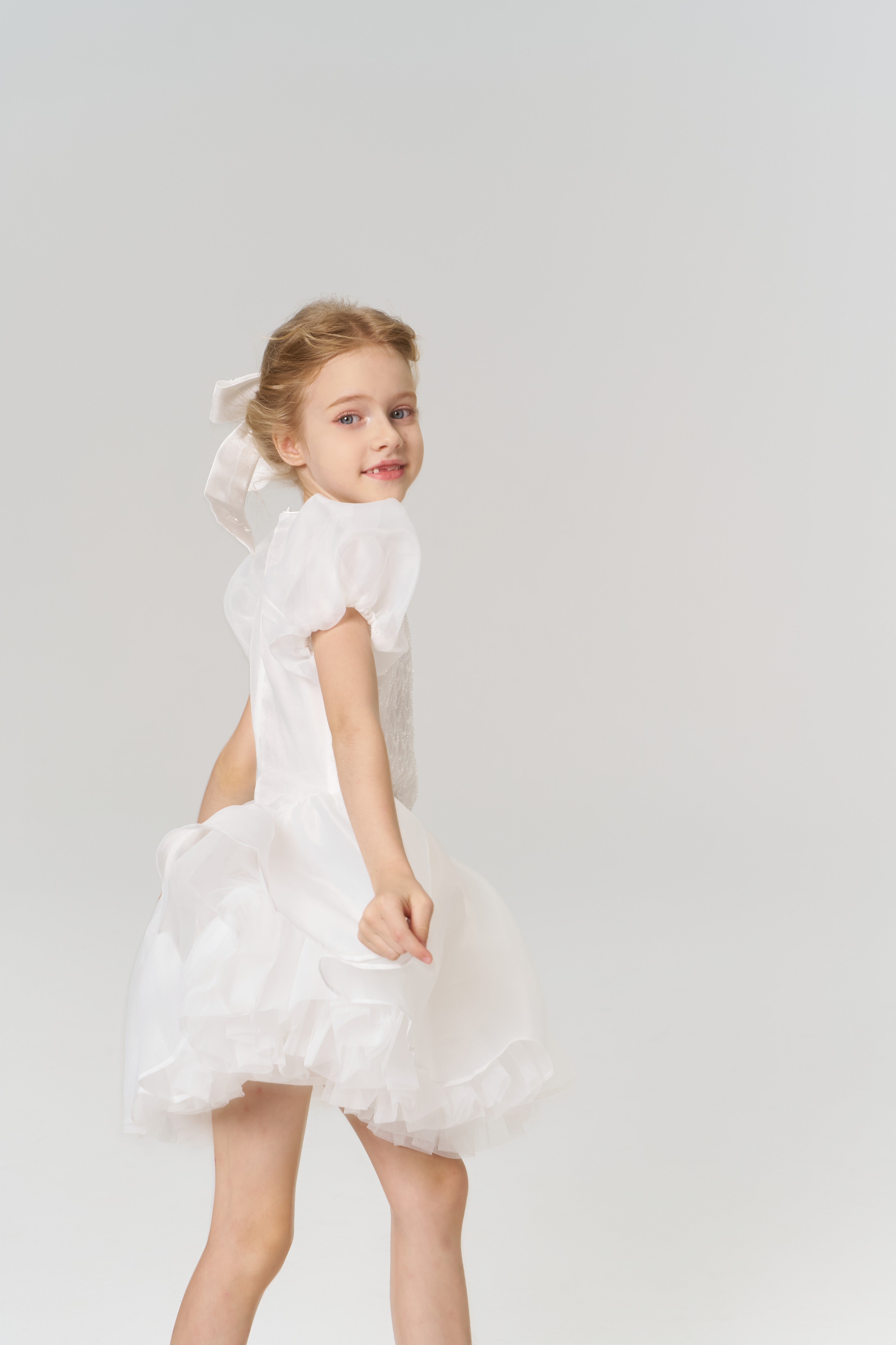 A young girl with a beaming smile wears the KIKI and JOJO Girls Hand-Beaded Pearl Satin Bow Hair Clip, pairing it beautifully with her white puffed-sleeve dress against a simple, light background.
