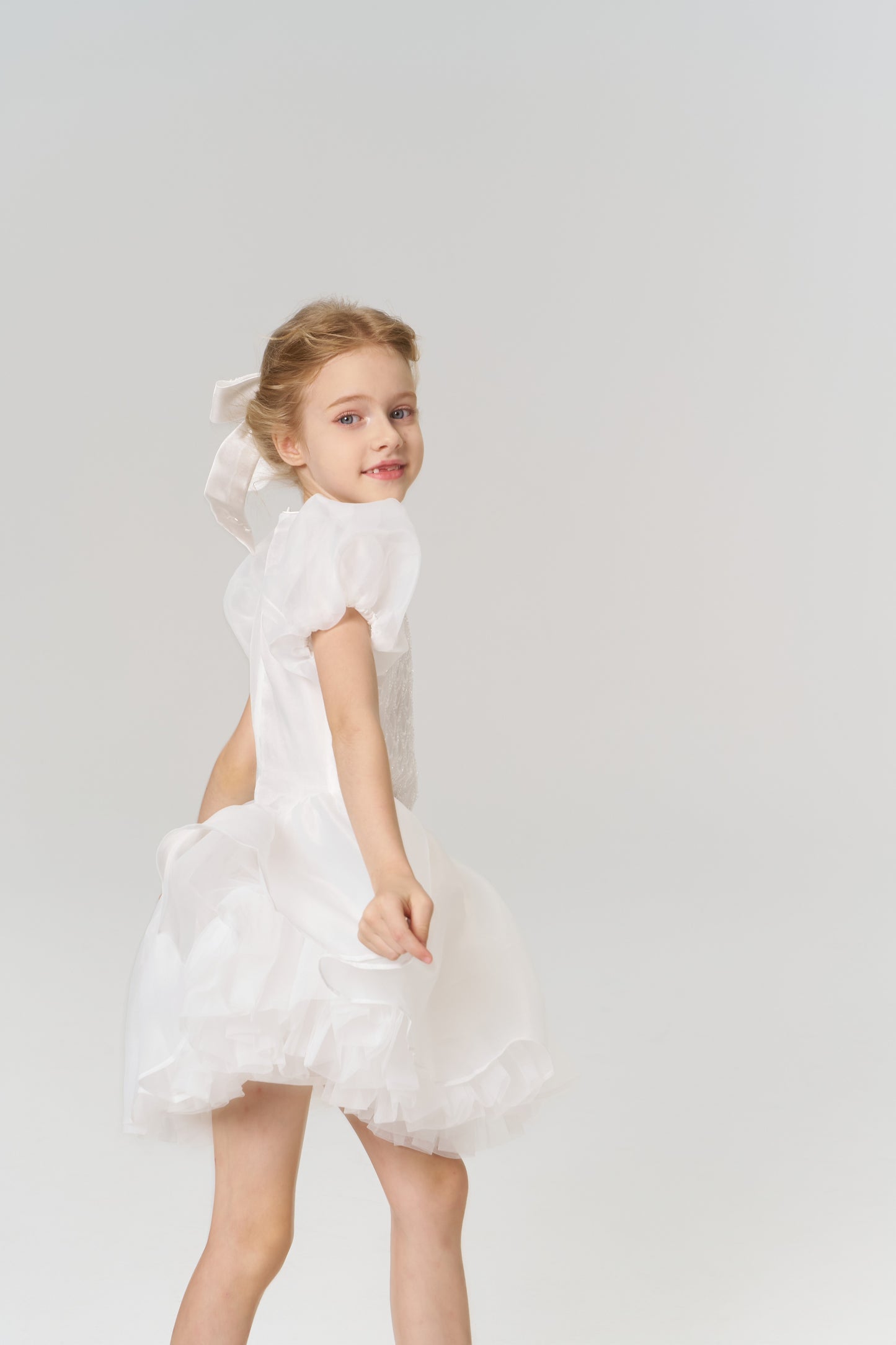 A young girl with a beaming smile wears the KIKI and JOJO Girls Hand-Beaded Pearl Satin Bow Hair Clip, pairing it beautifully with her white puffed-sleeve dress against a simple, light background.