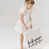 A young flower girl in a white dress with a satin bow wears the Girls Hand-Beaded Pearl Satin Bow Hair Clip by KIKI and JOJO, holding a large white shopping bag with "kikijapa" written in black script.